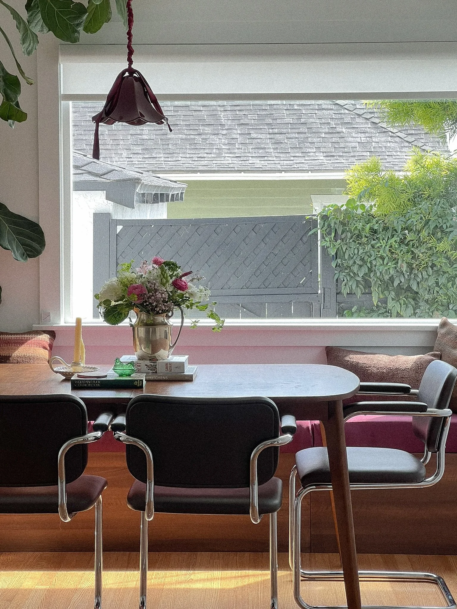 A view of a dining room with black chairs around a wooden table. On the table, there is a silver vase with flowers, books, and a candle. There is a large window with a view of a backyard fence, shrubs, and part of a house roof outside. A hanging lamp is above the table and some indoor plants are nearby.