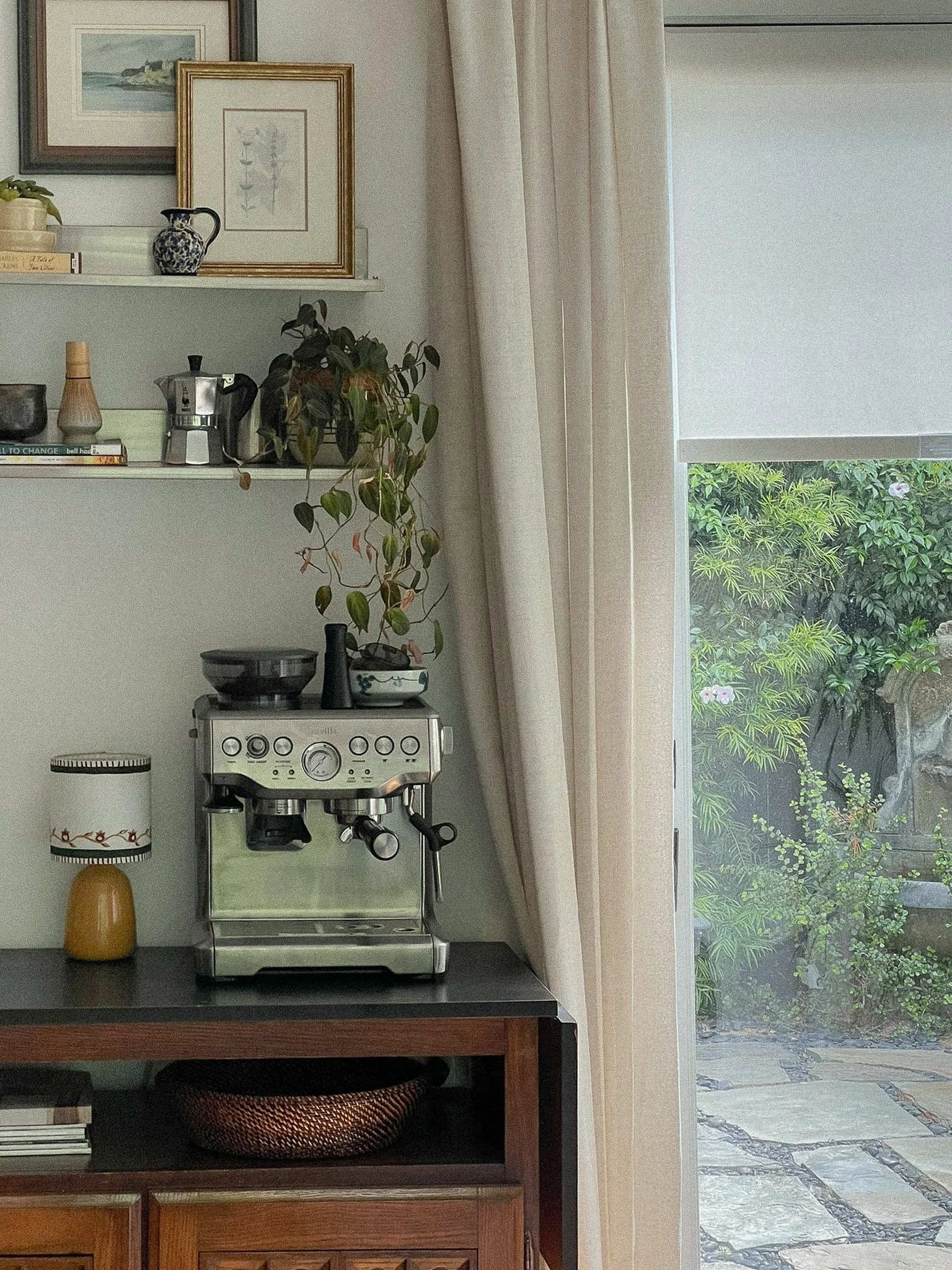 Interior view of a kitchen with a stove, a countertop, a shelf with framed pictures and decorative items, a potted plant, a lamp, and a large window with curtains showing a garden outside.