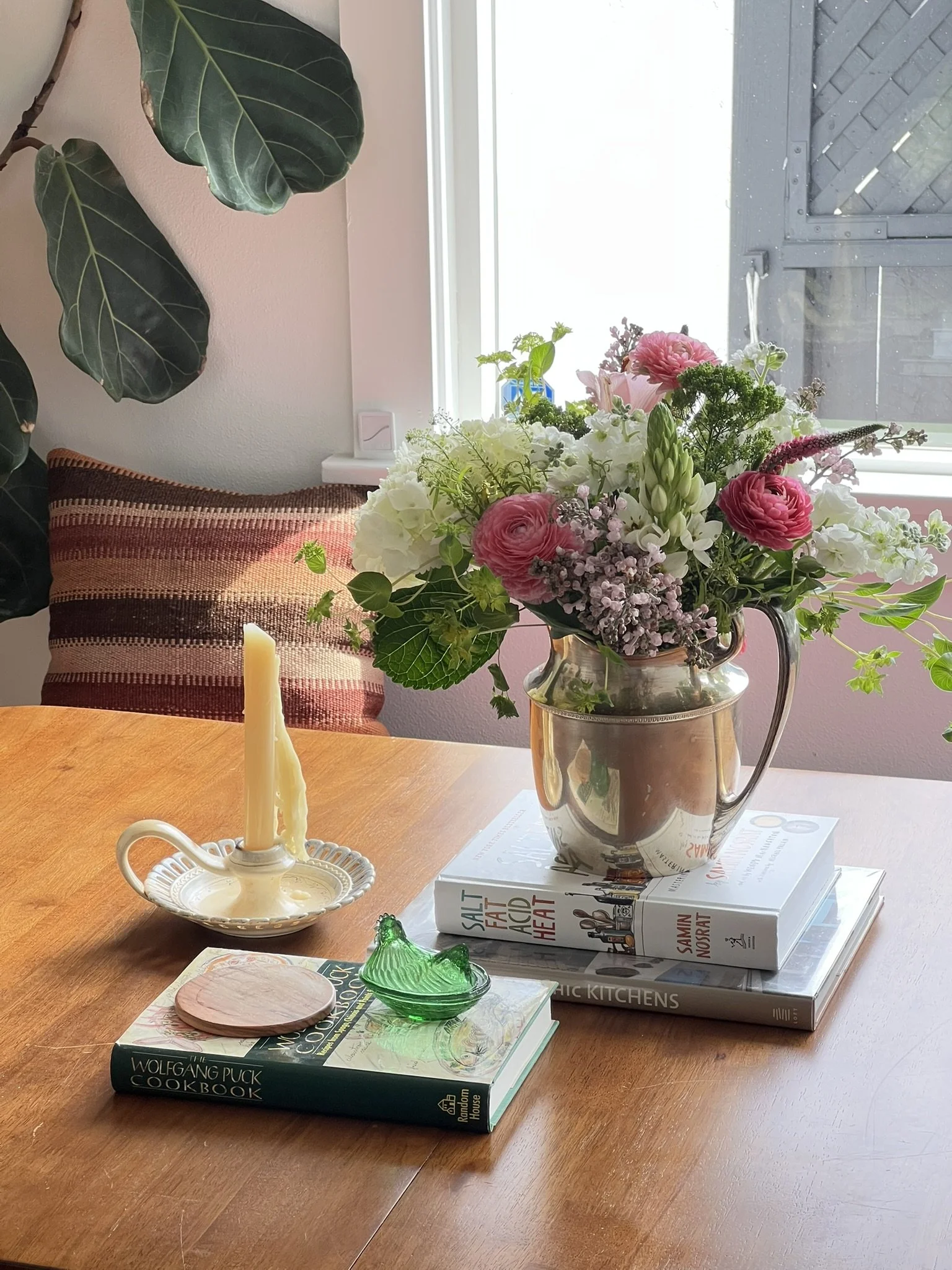 A wooden dining table with a bouquet of flowers in a glass jar, a stack of cookbooks, a green glass object, a candle in a holder, a small coaster, and some magazines, with a window in the background.