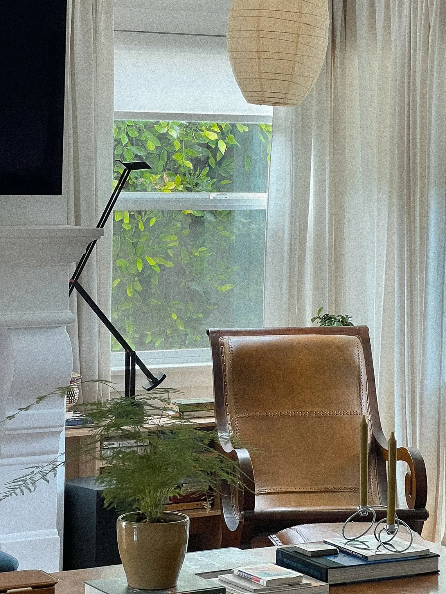 A cozy room with a large window showing greenery outside, a tan upholstered chair, a potted plant, books and papers on a desk, a black desk lamp, and a paper lantern hanging from the ceiling.