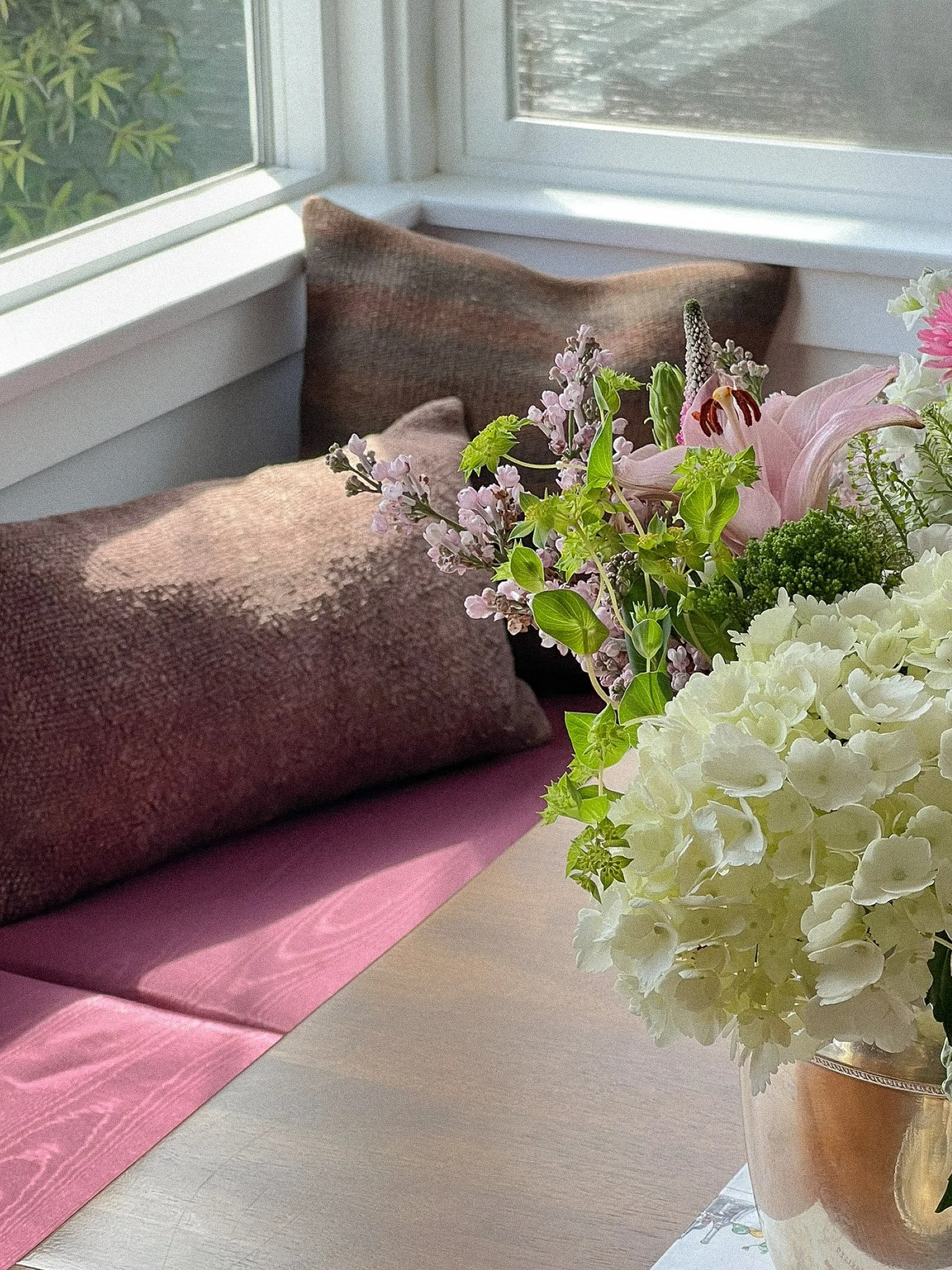 A bouquet of pink, white, and green flowers in a brown vase on a table next to pink and brown pillows by a window.