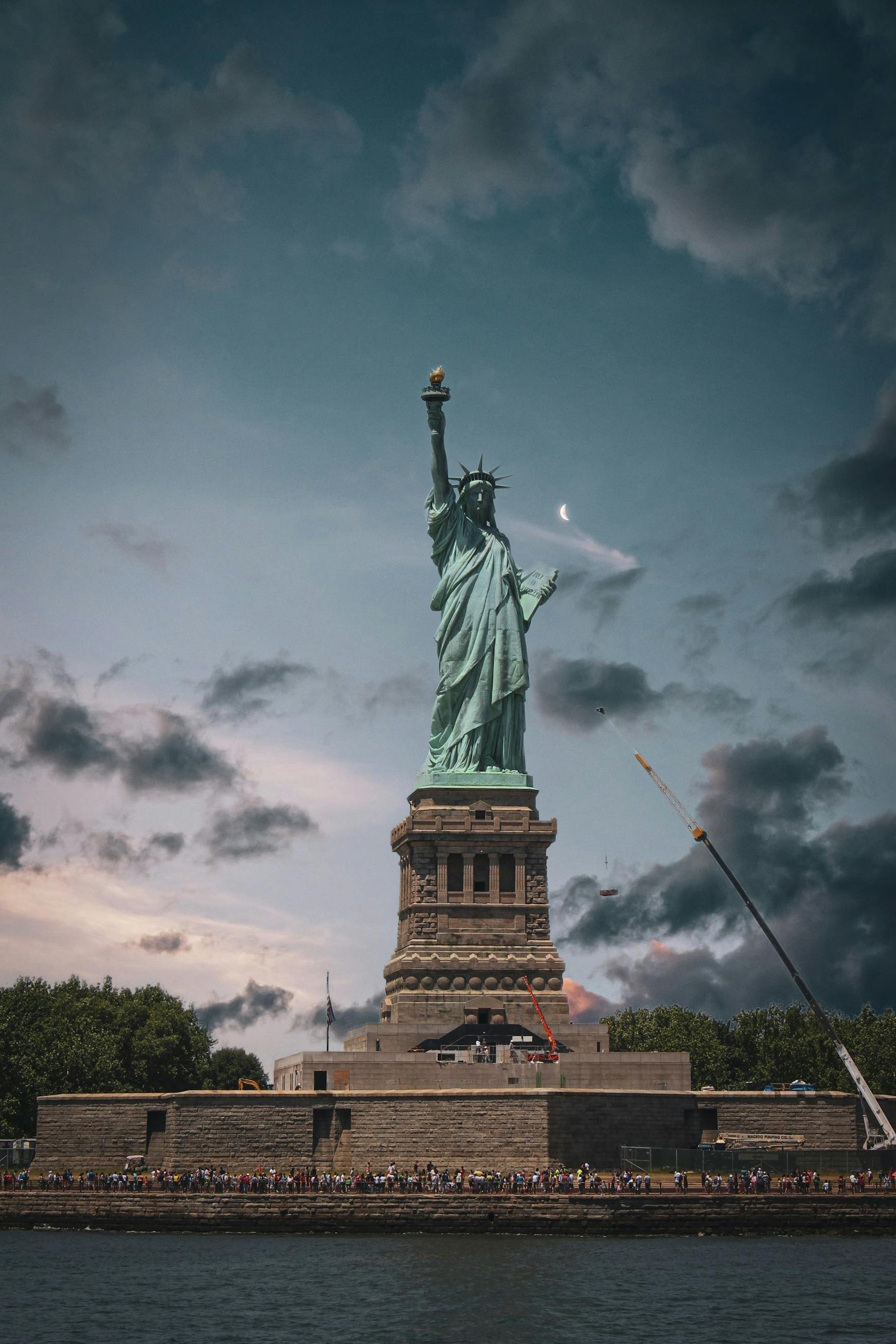 Photo of the Statue of Liberty under a cloudy sky with a crescent moon, a crowd of people at the base, and cranes nearby.
