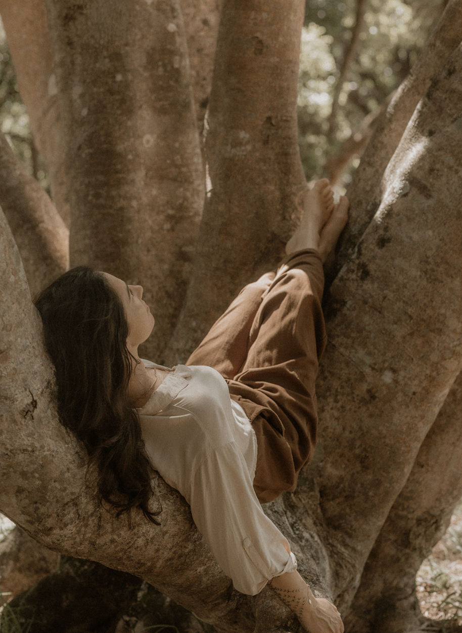 Woman lying on a large tree branch in a relaxed position, surrounded by tree trunks and leaves, with sunlight filtering through the foliage.