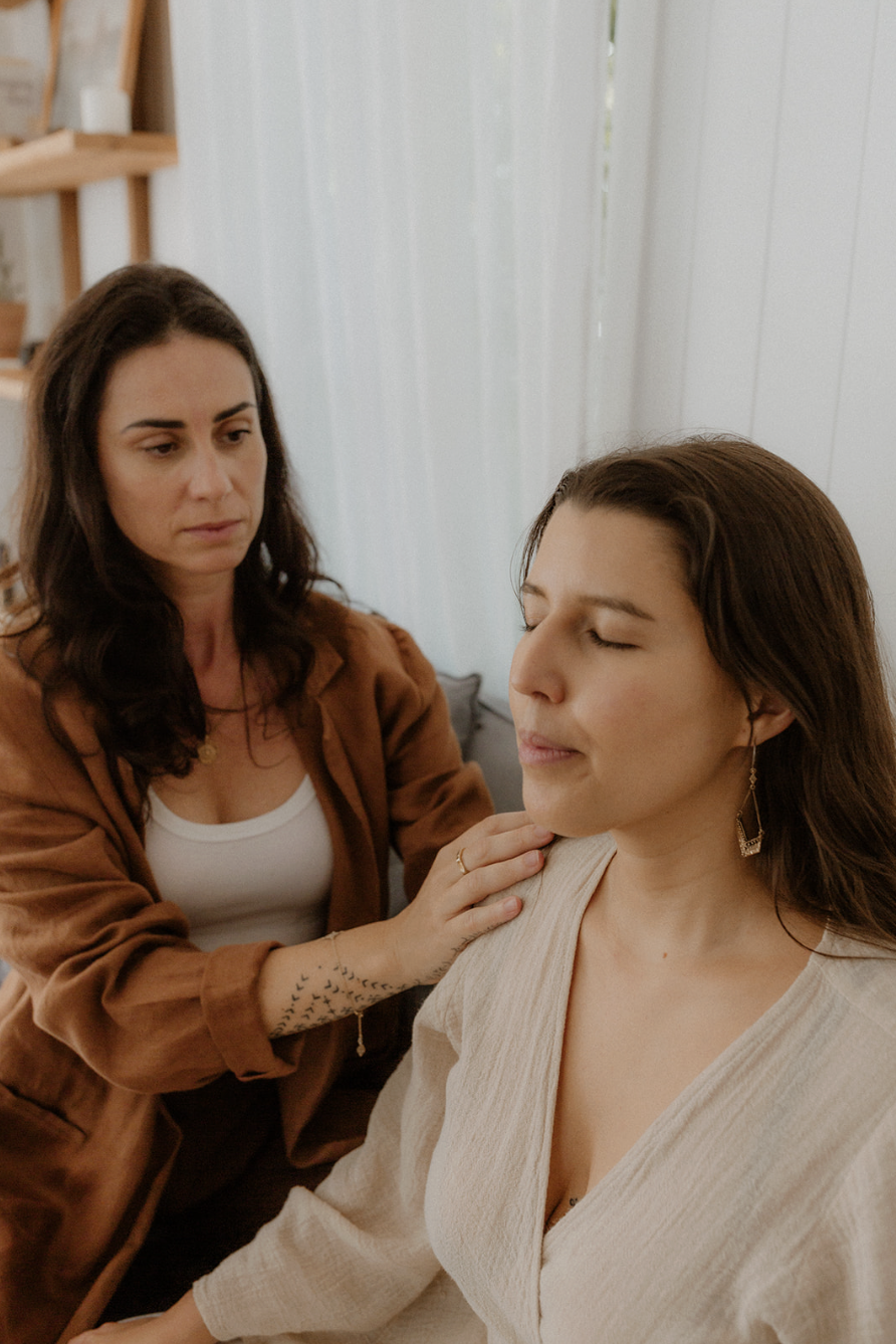 A woman with brown hair and earrings receives a gentle touch on her shoulder from another woman with dark hair, wearing a brown jacket, inside a room with white wooden walls.