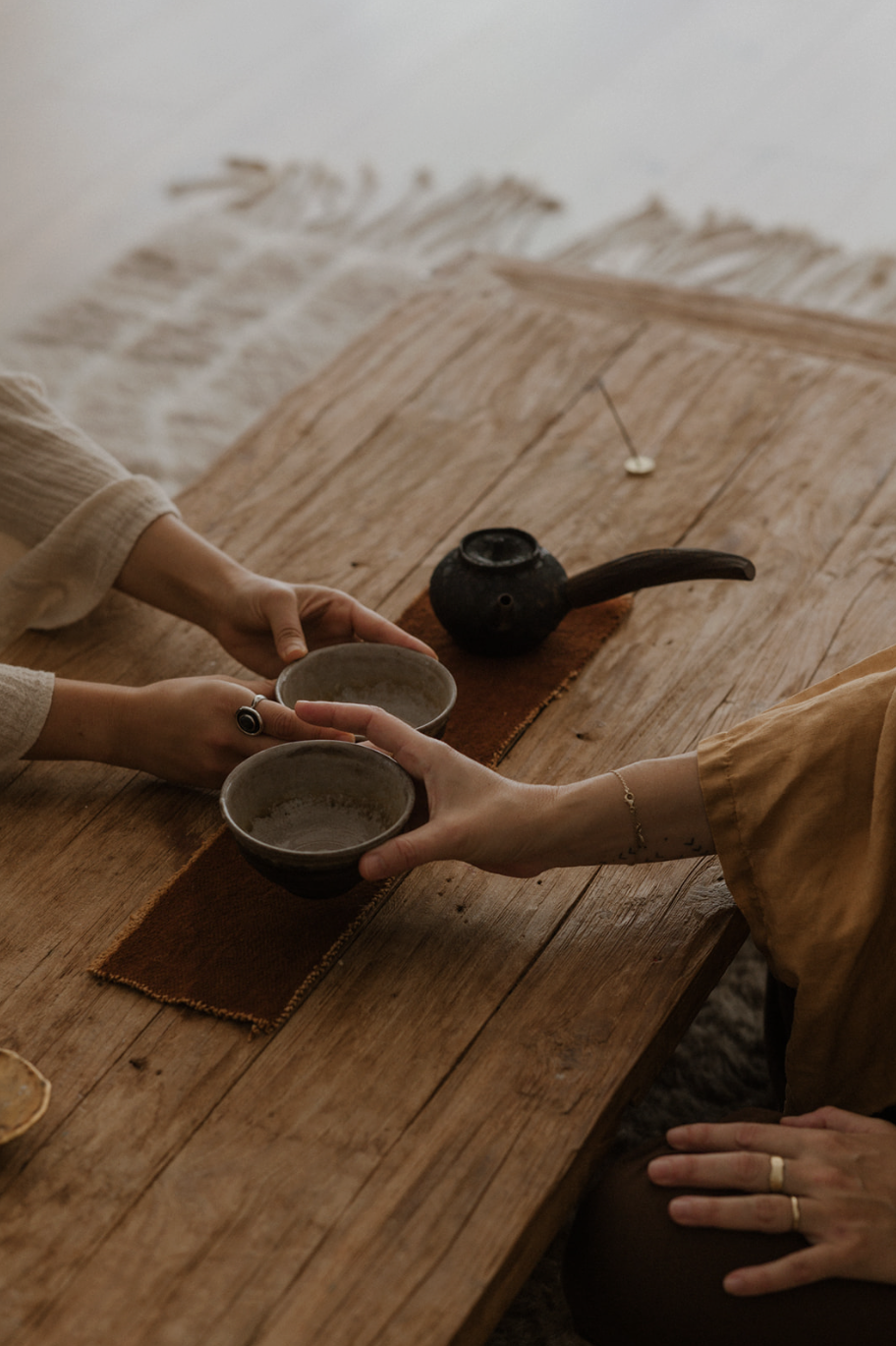 Two people exchanging ceramic cups over a wooden table with a small black teapot in the background.