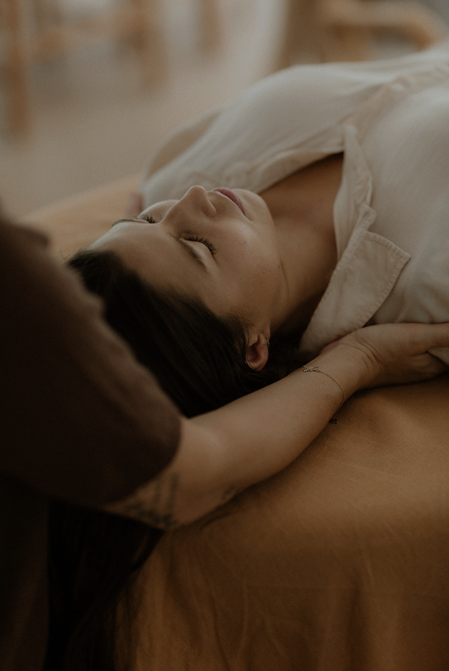 A woman lying on her back with eyes closed, resting on a bed, with a person beside her gently holding her head and arm.