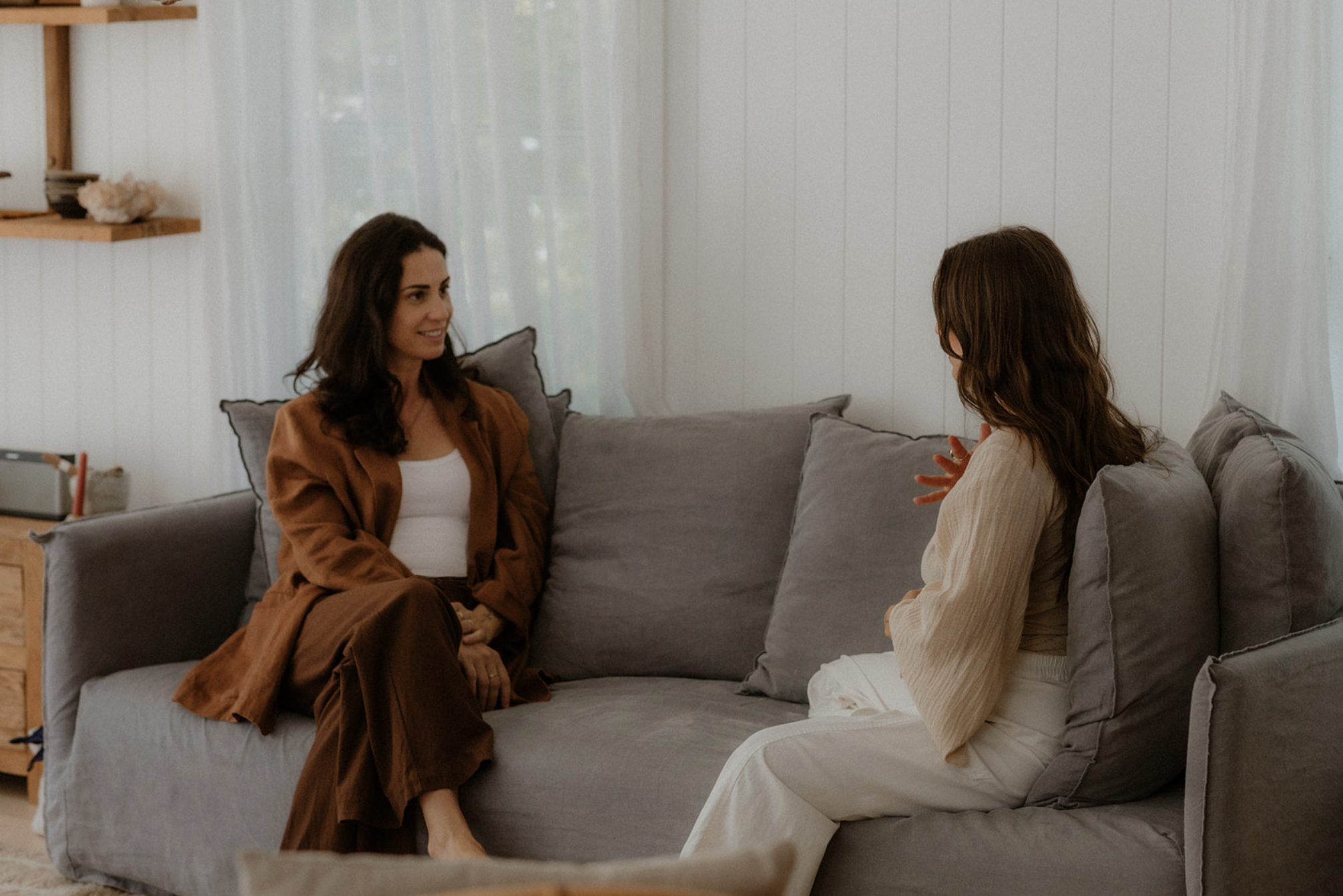 Two women engaging in a conversation on a gray sofa in a cozy room with white panel walls and sheer curtains.