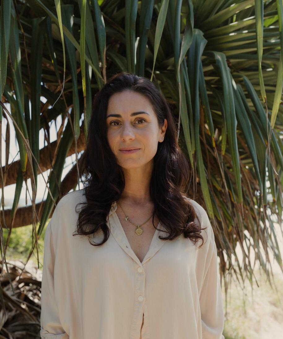 A woman with dark hair and a light-colored blouse standing outdoors in front of a large, leafy tree.