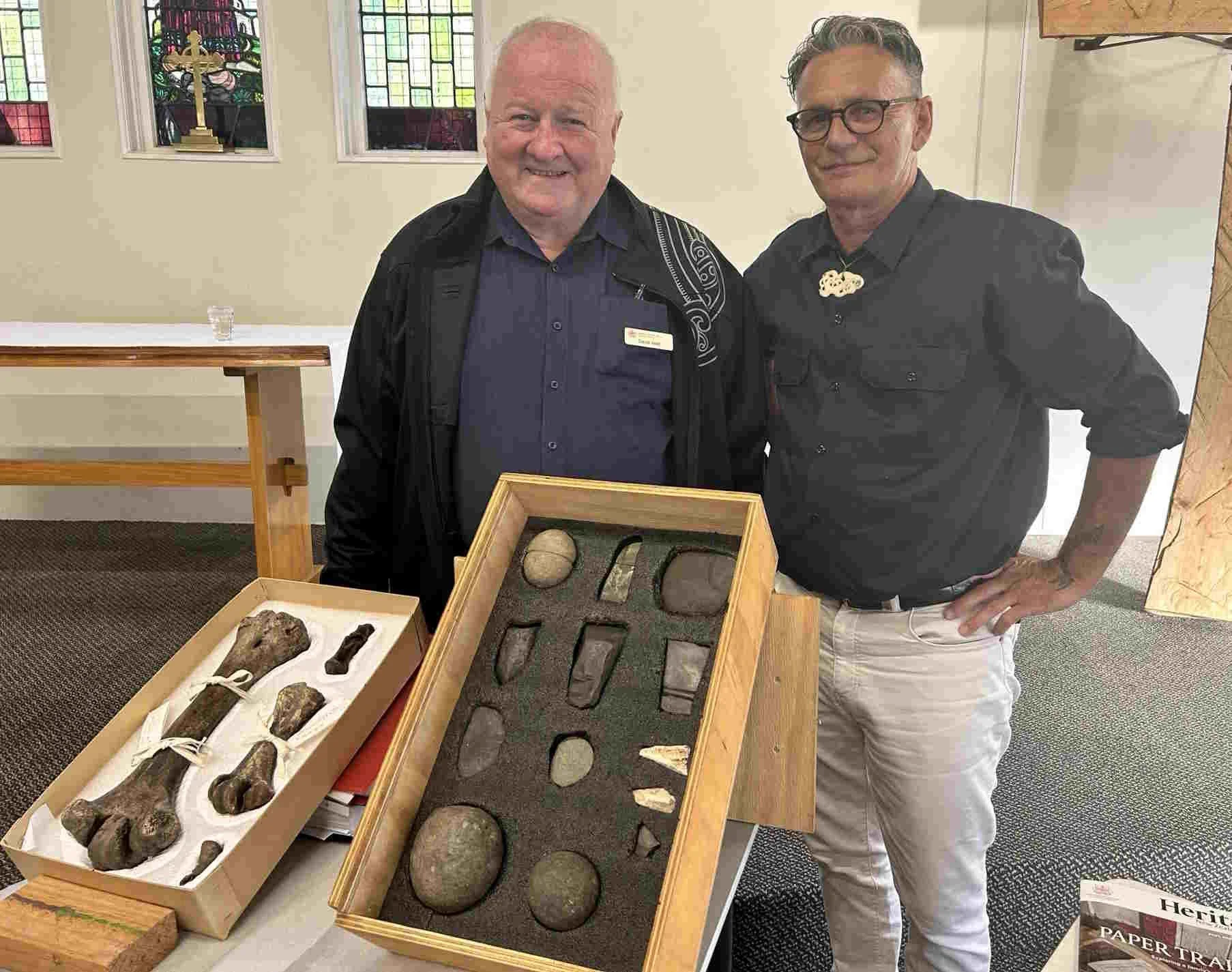 Two men standing behind tables displaying various archaeological artifacts, including stones and tools, inside a room with stained glass windows.