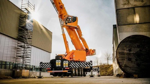 A large orange crane parked on a construction site between industrial buildings, with a cloudy sky overhead.