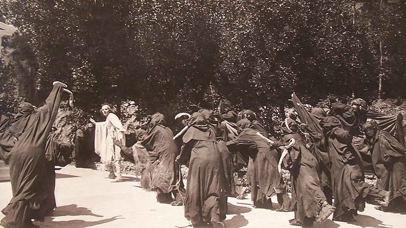 A group of hooded figures in dark robes and holding snakes threaten a young man in white.