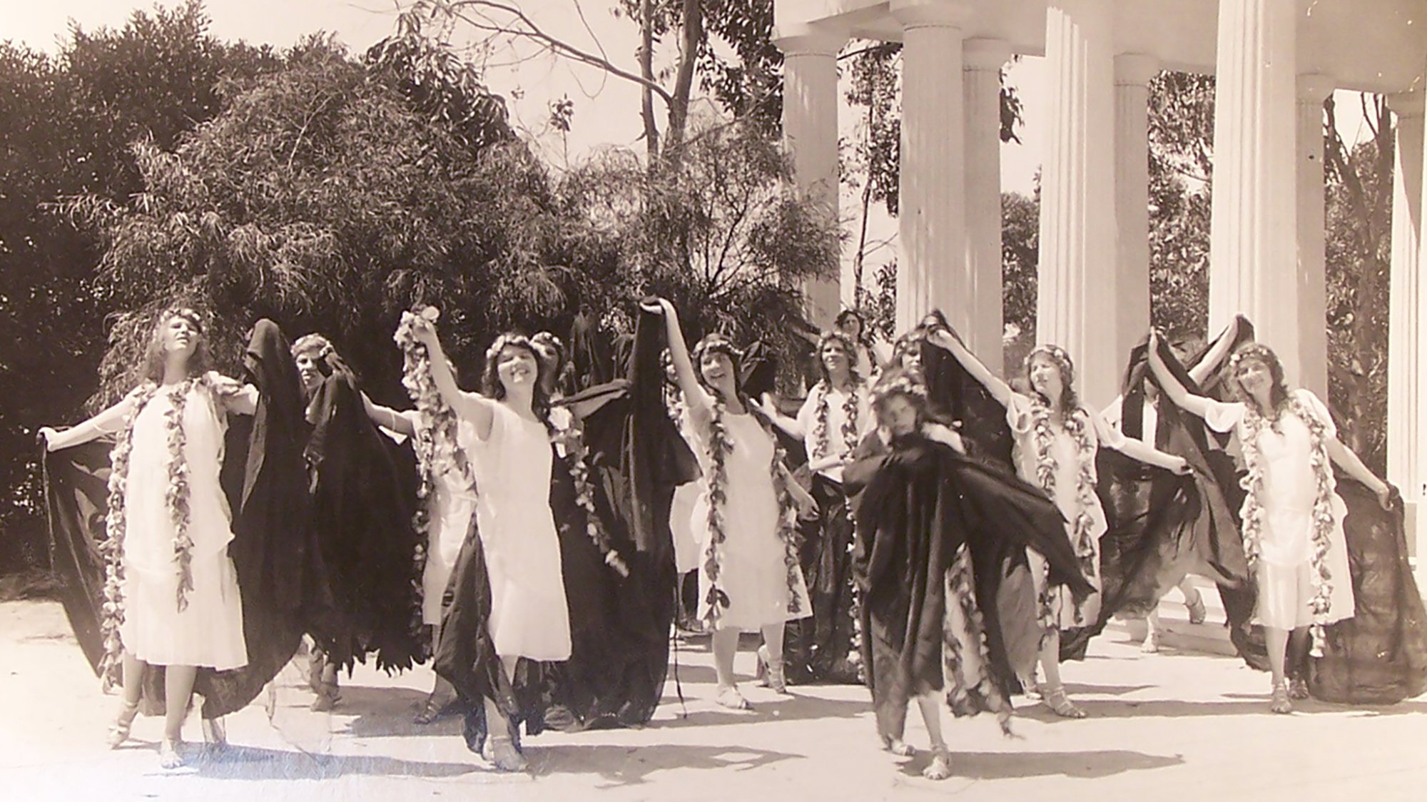 Young women throwing off dark, shredded robes to reveal fresh white robes and flower garlands underneath.