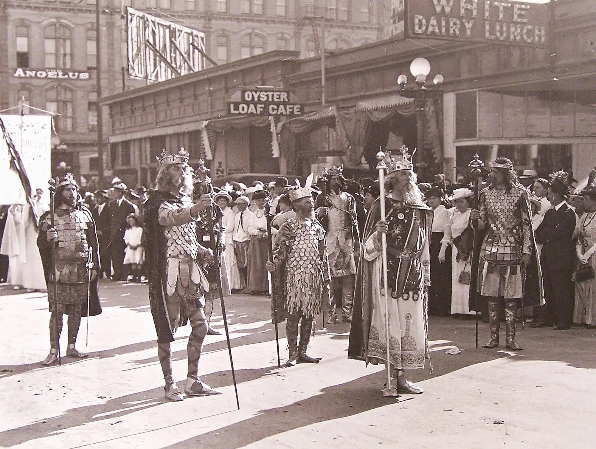 Seven actors dressed in the attire of medeival kings walking down the street in San Diego in the year 1914.