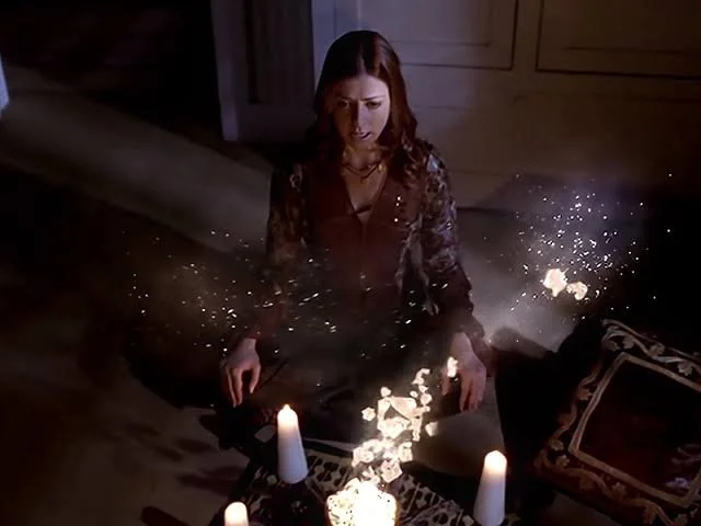 A redheaded young woman, Willow, sits cross-legged on the floor in front of lit candles, casting a shimmering spell.