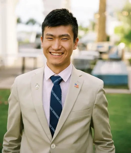 Young man in a light gray suit with a blue tie, smiling outdoors.