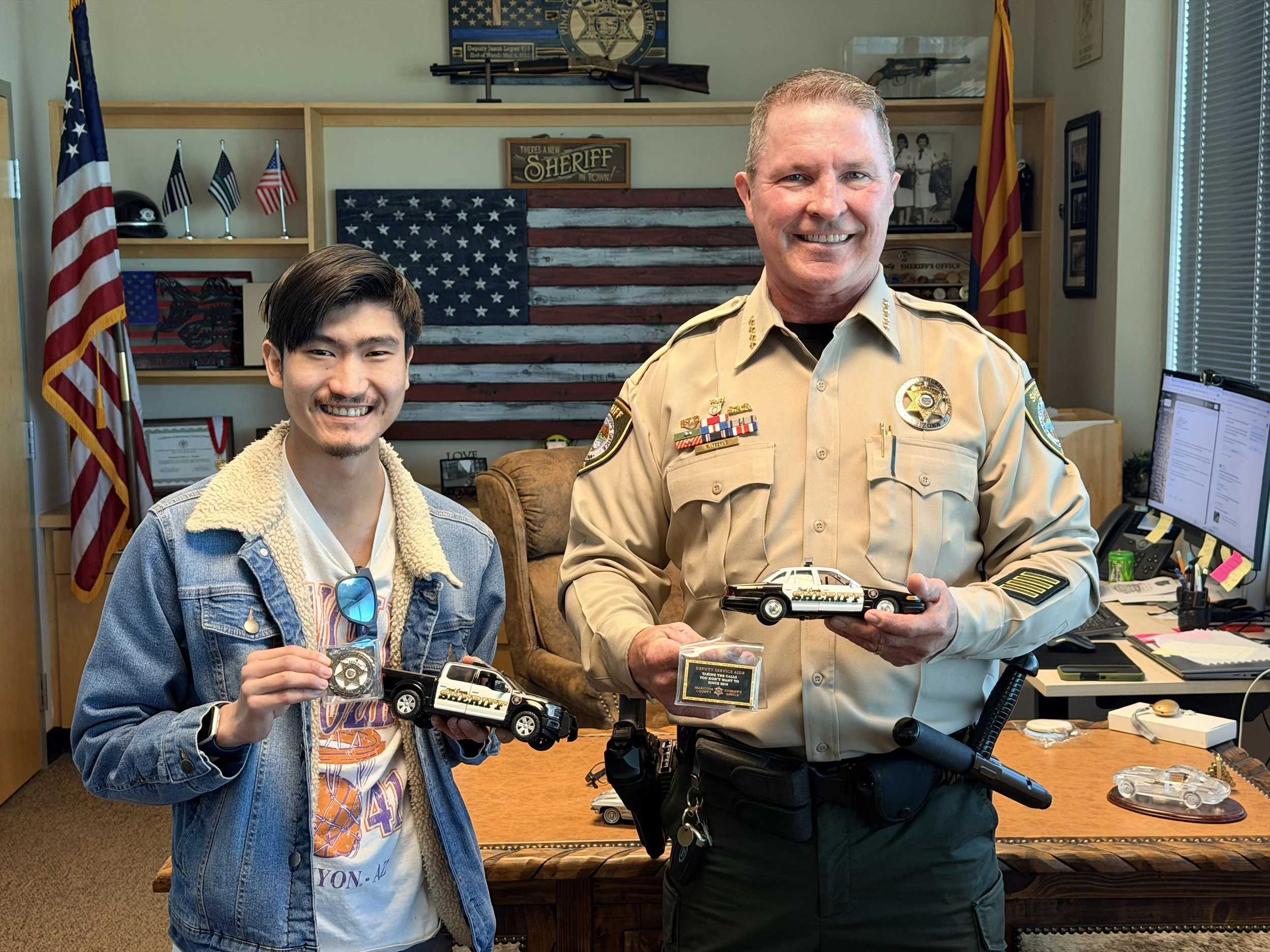 A young man and a police officer standing indoors, holding small sheriff badge and toy police car model. They are smiling. The room decor includes American flags, a sheriff's badge, an American flag-themed wall art, and police memorabilia.