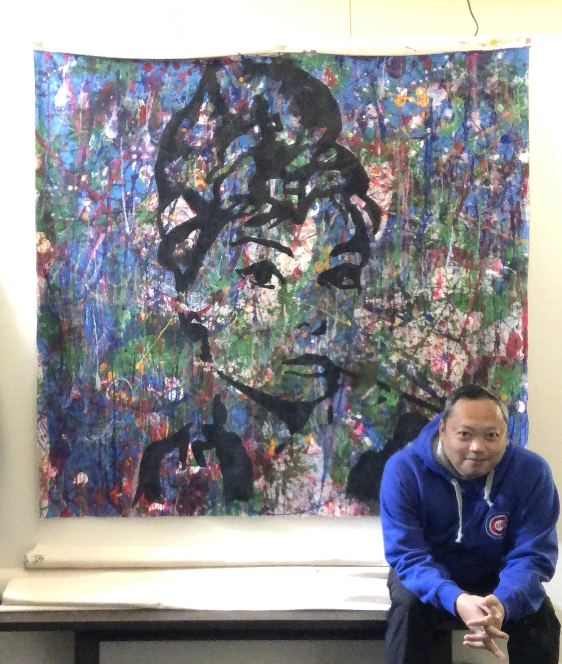 A man in a blue Chicago Cubs jacket crouches in front of a colorful abstract painting with a black tree silhouette on it.