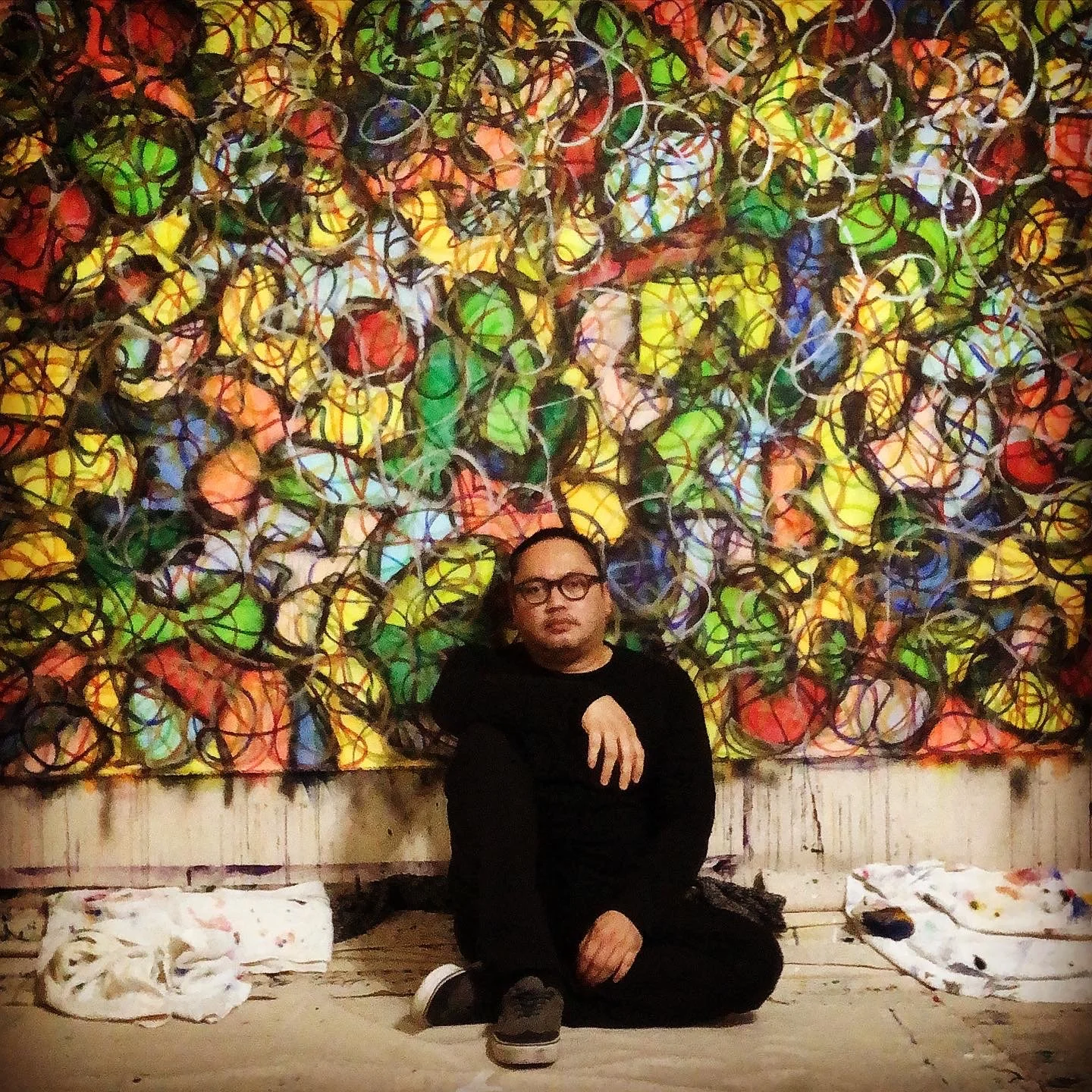 A person with glasses wearing a black shirt is sitting on the floor in front of a colorful, abstract mural of interconnected circles and lines. They are seated cross-legged with one hand resting on their knee, and the background features a wall covered with this vibrant artwork.