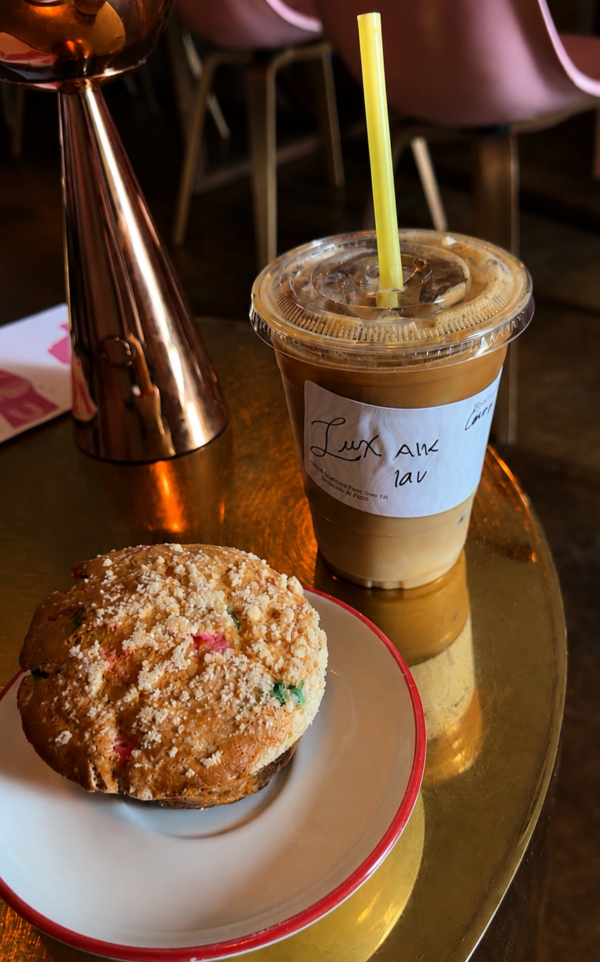 A large iced coffee drink with a yellow straw on a shiny, gold-colored table, next to a decorated cookie on a small white plate with a red rim, in a cozy cafe setting.