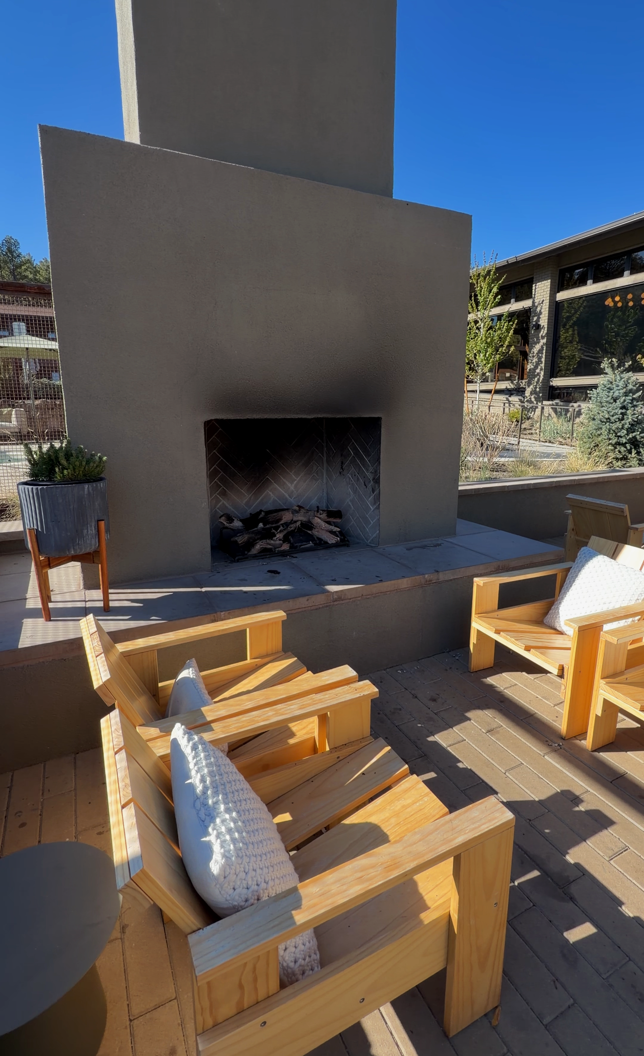 Outdoor patio with wooden chairs seats with white cushions and pillows, a building with large windows, a fireplace with wood logs, potted plants, and a clear blue sky.