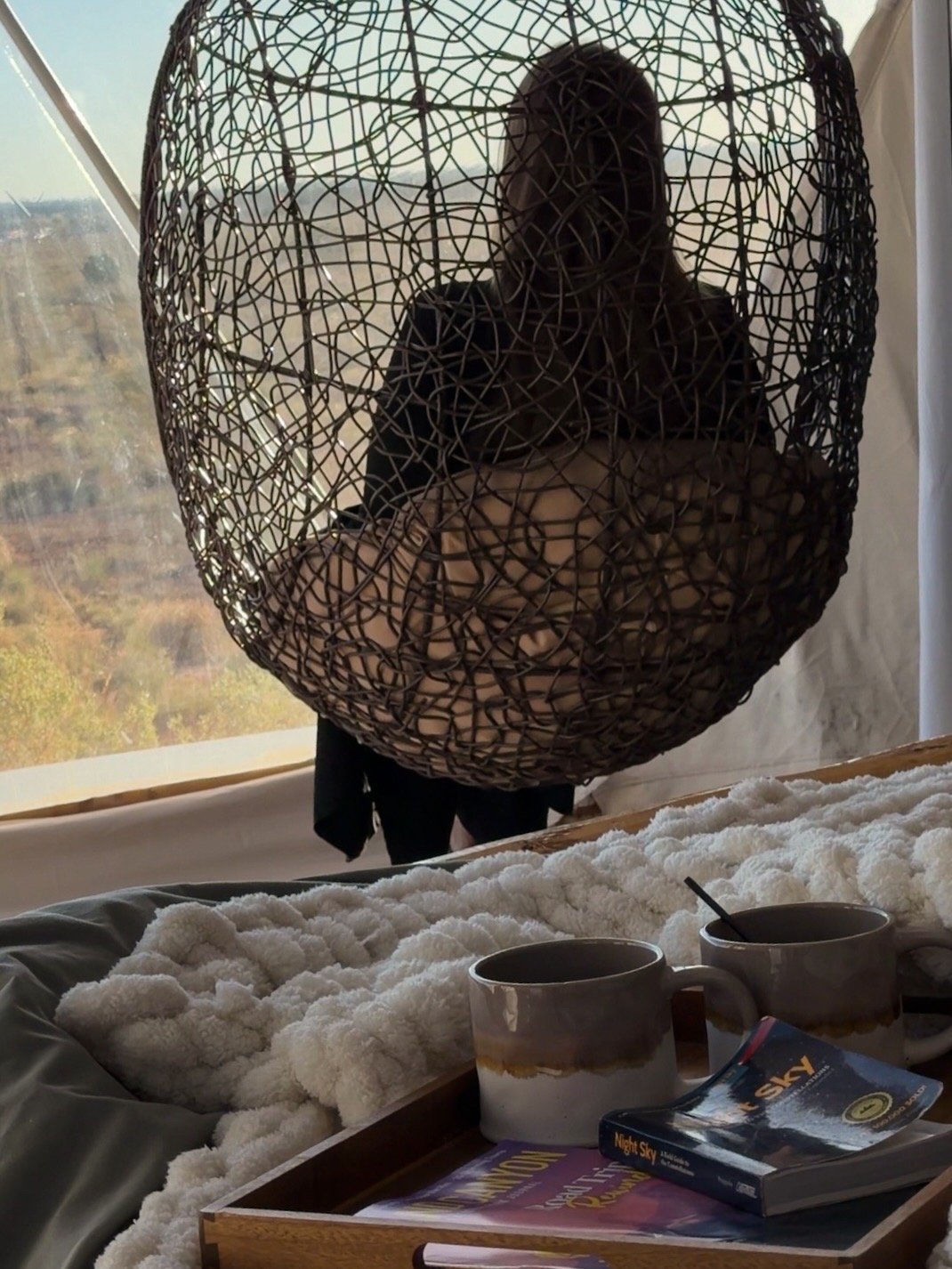 Person taking a photo in a decorative, woven, basket-style mirror reflection, with a bed, two coffee mugs, and a tray with a book and a spoon on a fuzzy blanket in the foreground.