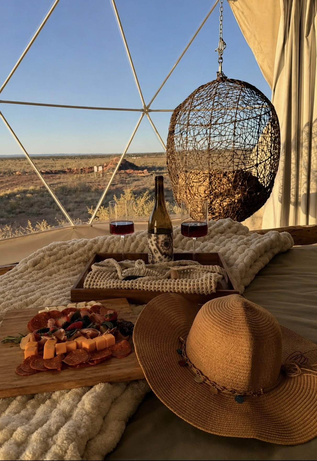 A cozy indoor scene featuring a tray of assorted cured meats and cheeses, two glasses of red wine, a wine bottle, a straw sun hat, and a platter of fruit, set on a plush blanket near a geodesic dome window with a desert landscape view.