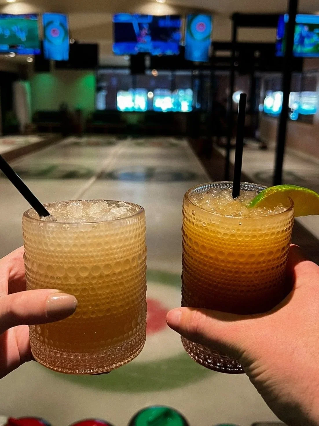 Two hands holding glasses of yellowish drinks with ice, black straws, and lime wedges, at a bowling alley with multiple television screens in the background.