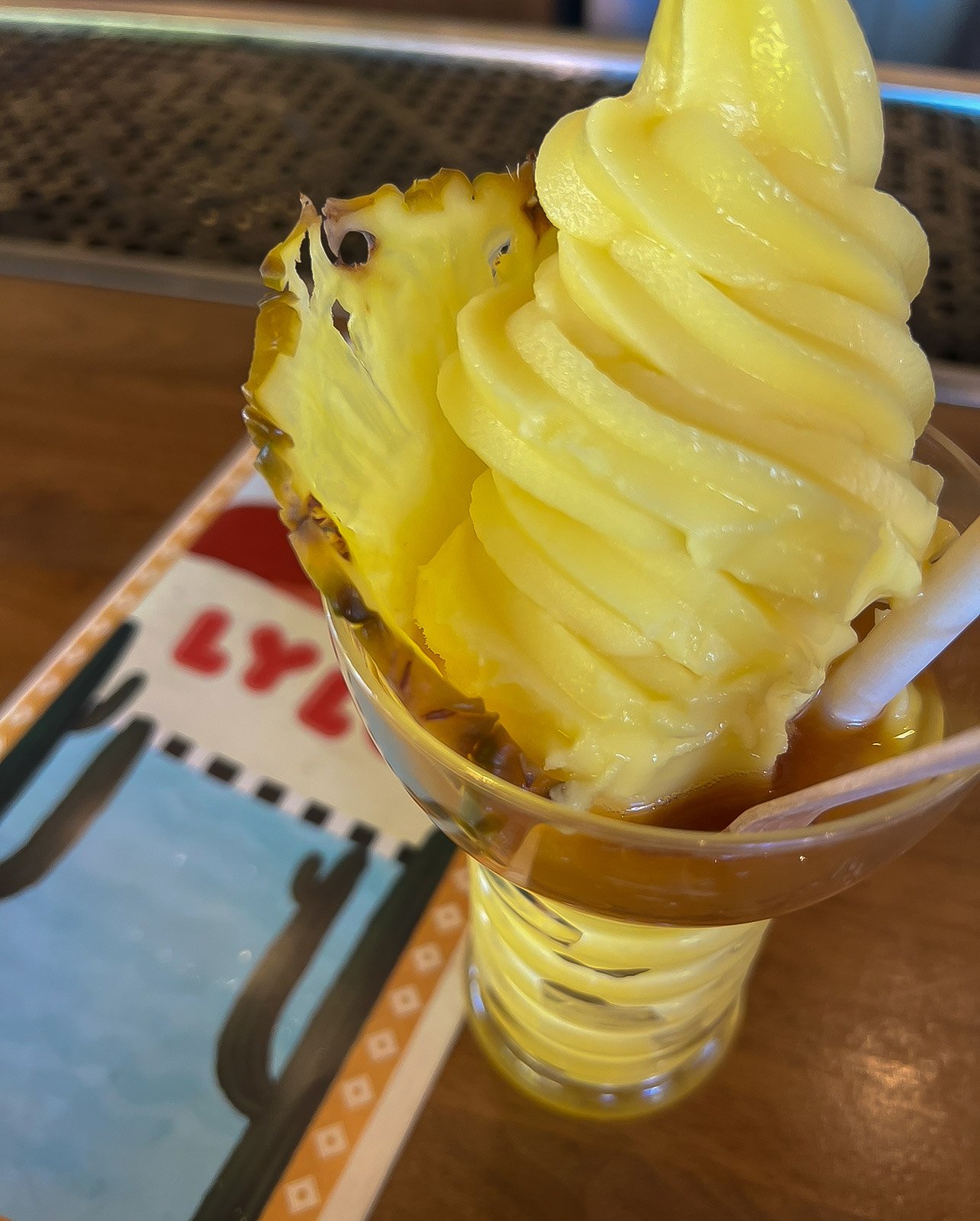 A tall glass of frozen pineapple mousse topped with a pineapple slice, with a straw in it, on a wooden table with a colorful paper placemat underneath.