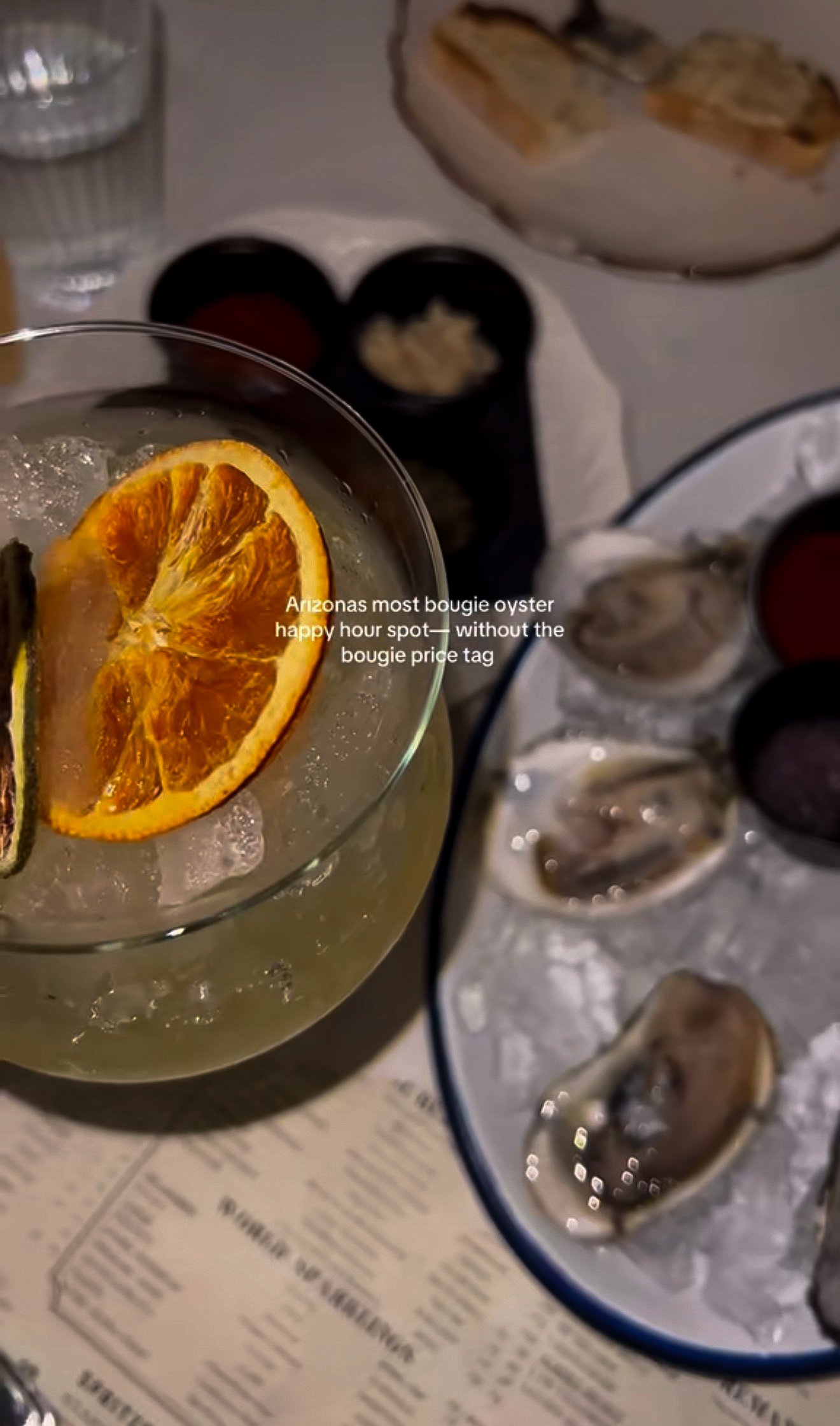 A glass with a citrus slice and ice, a plate with oysters, and a menu in the background.