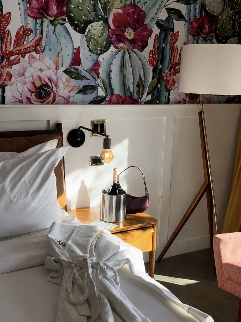 A cozy bedroom corner with a bed, white bedding, and a robe. There is a wooden nightstand with a bottle of champagne in an ice bucket and a purple handbag. A wall-mounted lamp is on, and a tall floor lamp with a white shade stands to the side. A colo