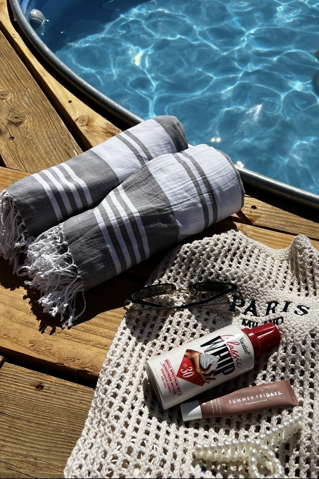 Towels, sunglasses, sunscreen, a pearl necklace, and a crocheted bag on a wooden deck by a swimming pool.