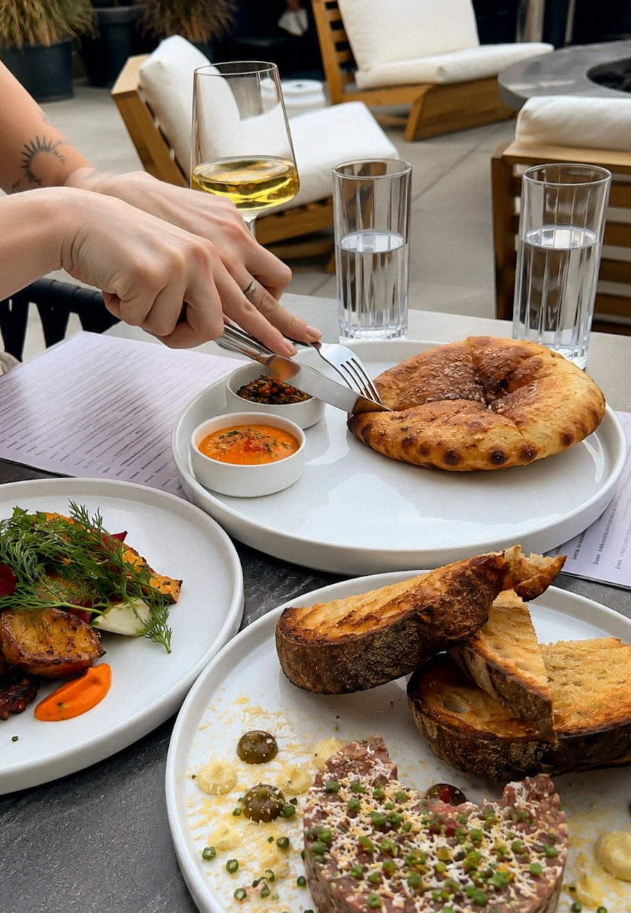 A person using a fork and knife to cut towards a piece of bread on a white plate at an outdoor dining table with various dishes, glasses of water, and a glass of white wine. The table has plates with appetizers and main courses, including grilled bread, a salad, and small bowls of sauce.