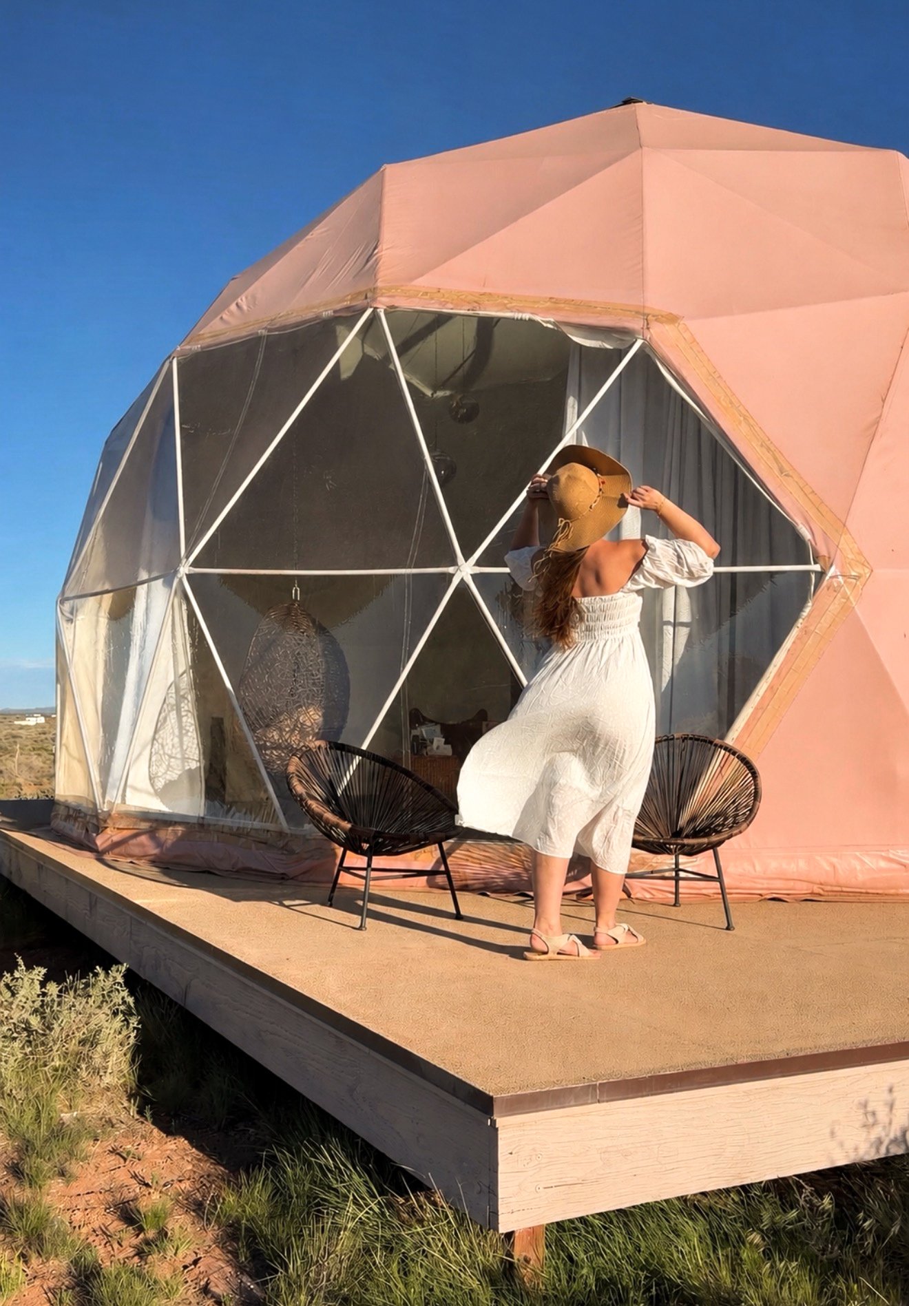 A woman wearing a white dress and a wide-brimmed hat stands on a wooden deck outside a geodesic dome with pink panels and a transparent section, in a desert landscape during sunset.