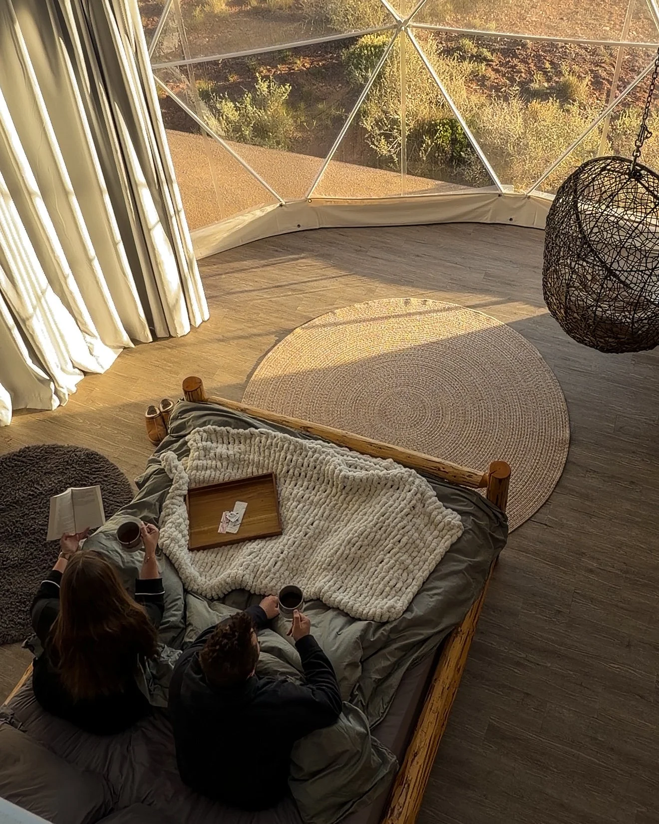 A cozy indoor space with a bed, a round rug, and two people sitting on the bed with cups, one holding a book, near large windows showing a view of trees and a hillside outside.