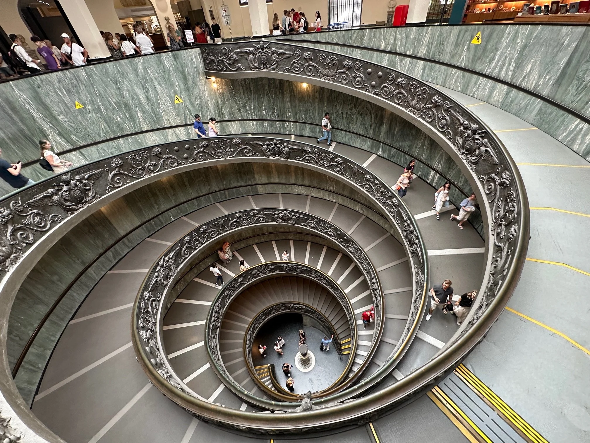 Spiral staircase viewed from above with decorative railing and people walking on the stairs and at the top level in a shopping mall.