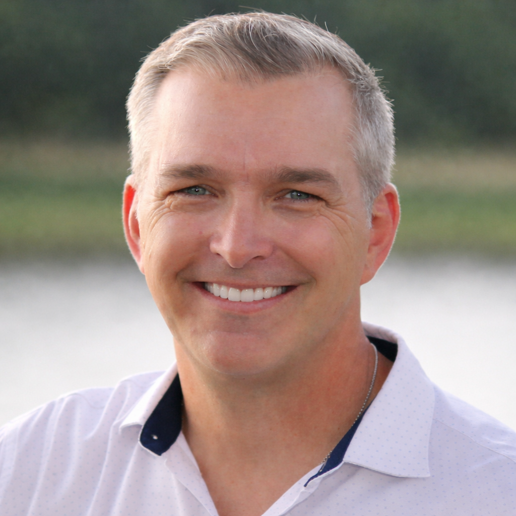 A smiling middle-aged man with short gray hair, blue eyes, wearing a white collared shirt with a dark trim, standing outdoors near a body of water with trees in the background.