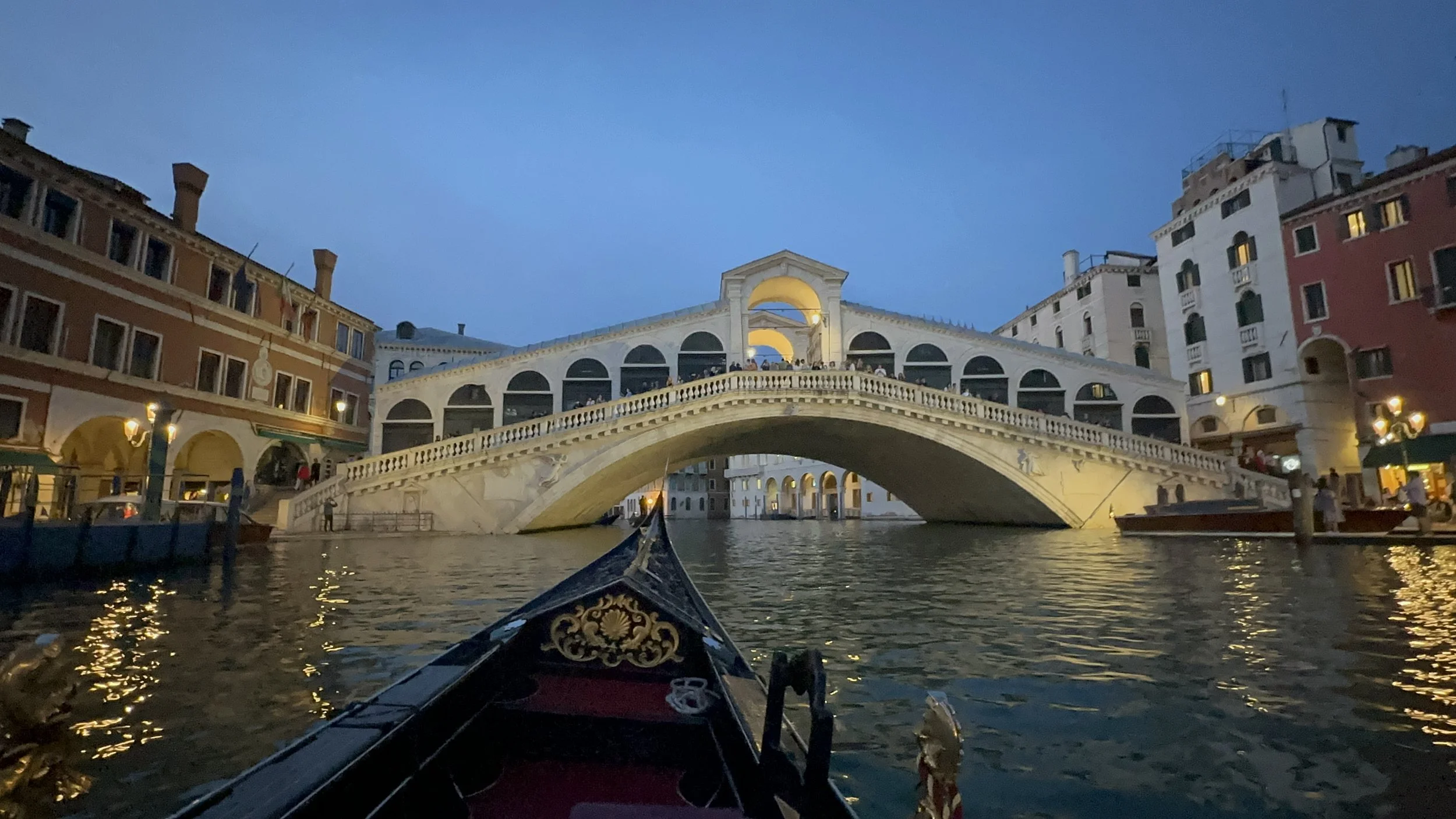 Photo taken from a boat on a canal showing the Rialto Bridge illuminated during dusk, with surrounding historic buildings and reflections on the water.