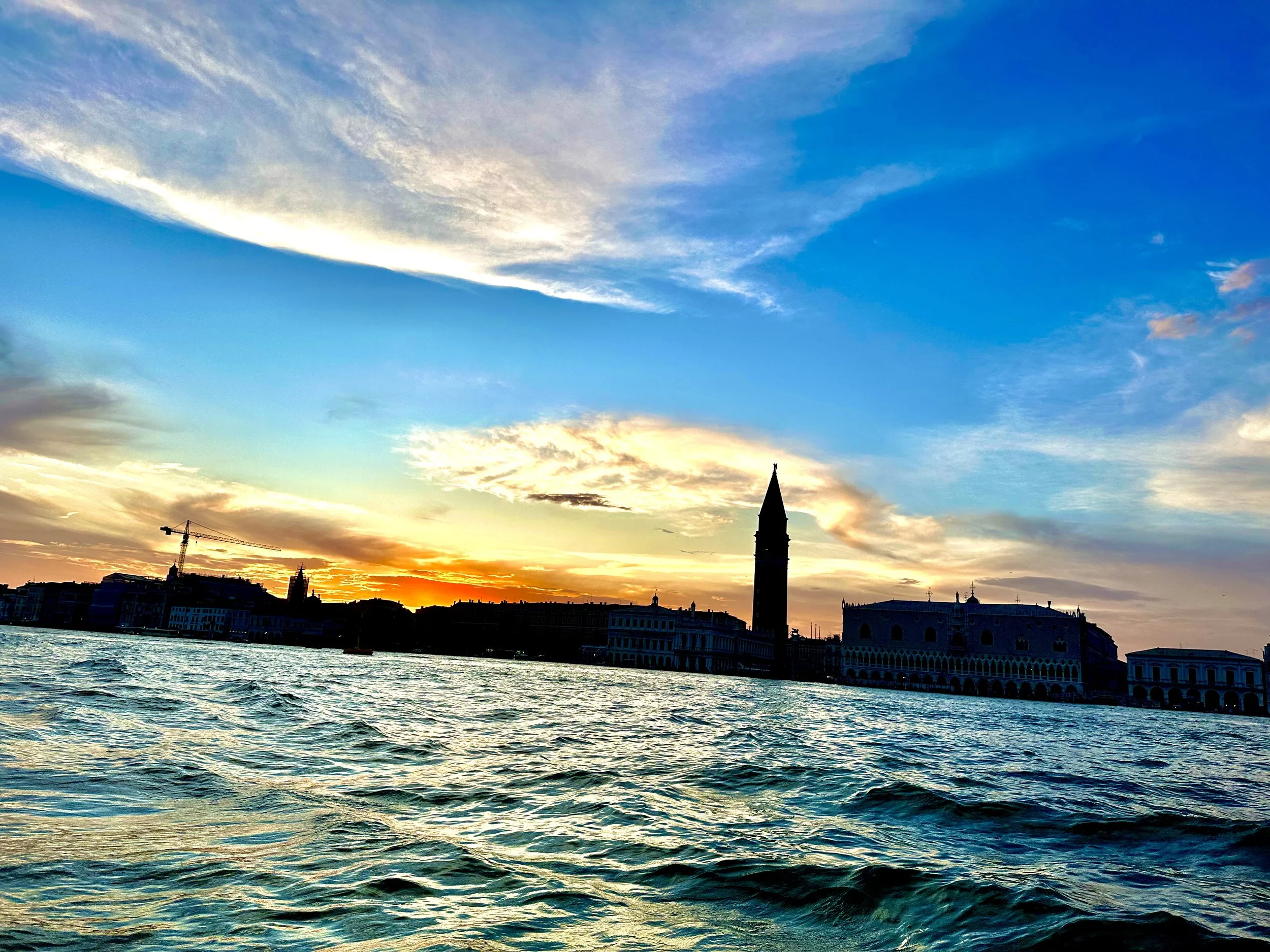 Sunset over Venice with water in the foreground and historic buildings and a tower silhouette in the background.