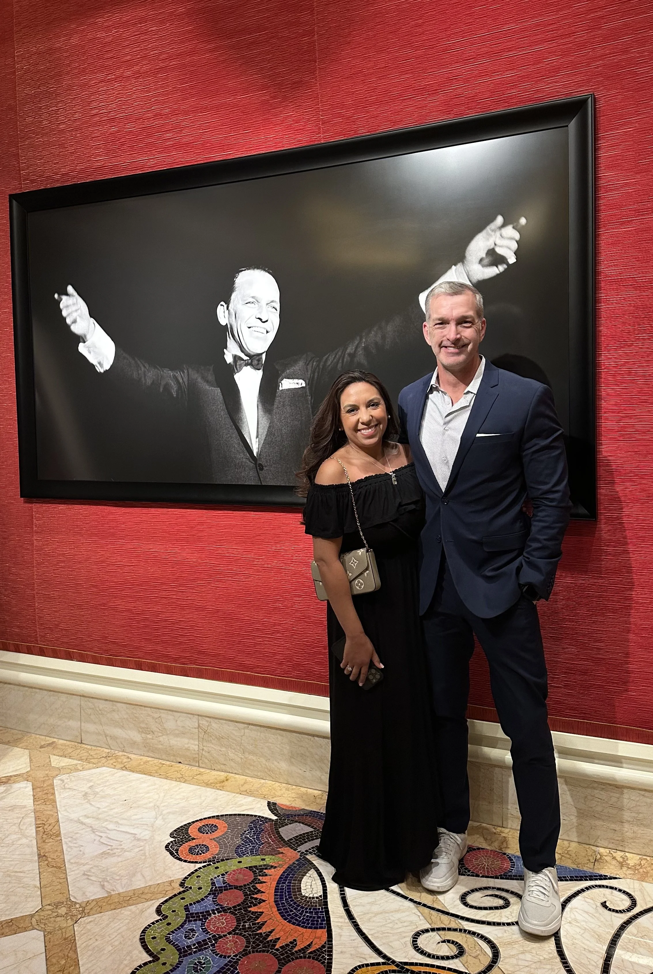 A man and woman standing together in front of a black and white portrait of a man in a tuxedo with arms outstretched, inside a room with red walls and decorative mosaic flooring.