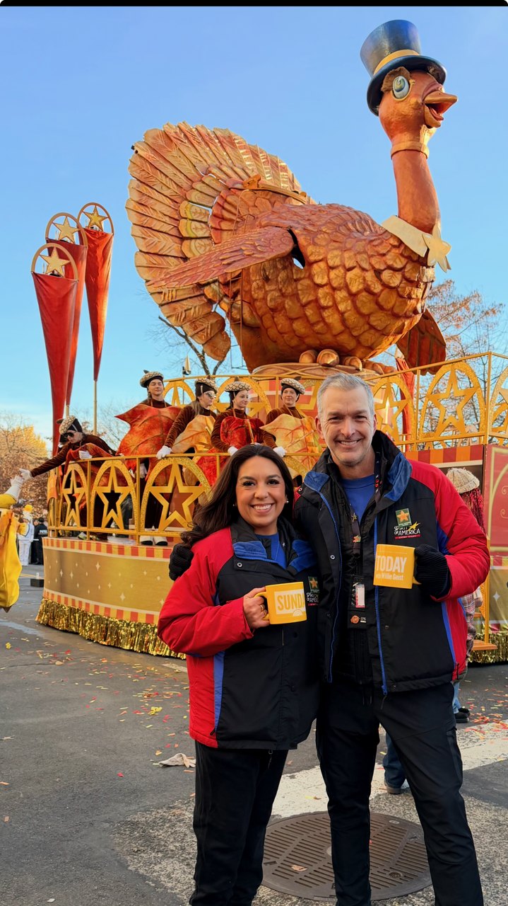 Two people smiling and holding mugs labeled 'SUNDAY' and 'TODAY' in front of a large parade float featuring a giant turkey wearing a top hat, with performers dressed in festive costumes. The float is decorated with stars and gold accents, and the background shows clear blue sky and autumn trees.
