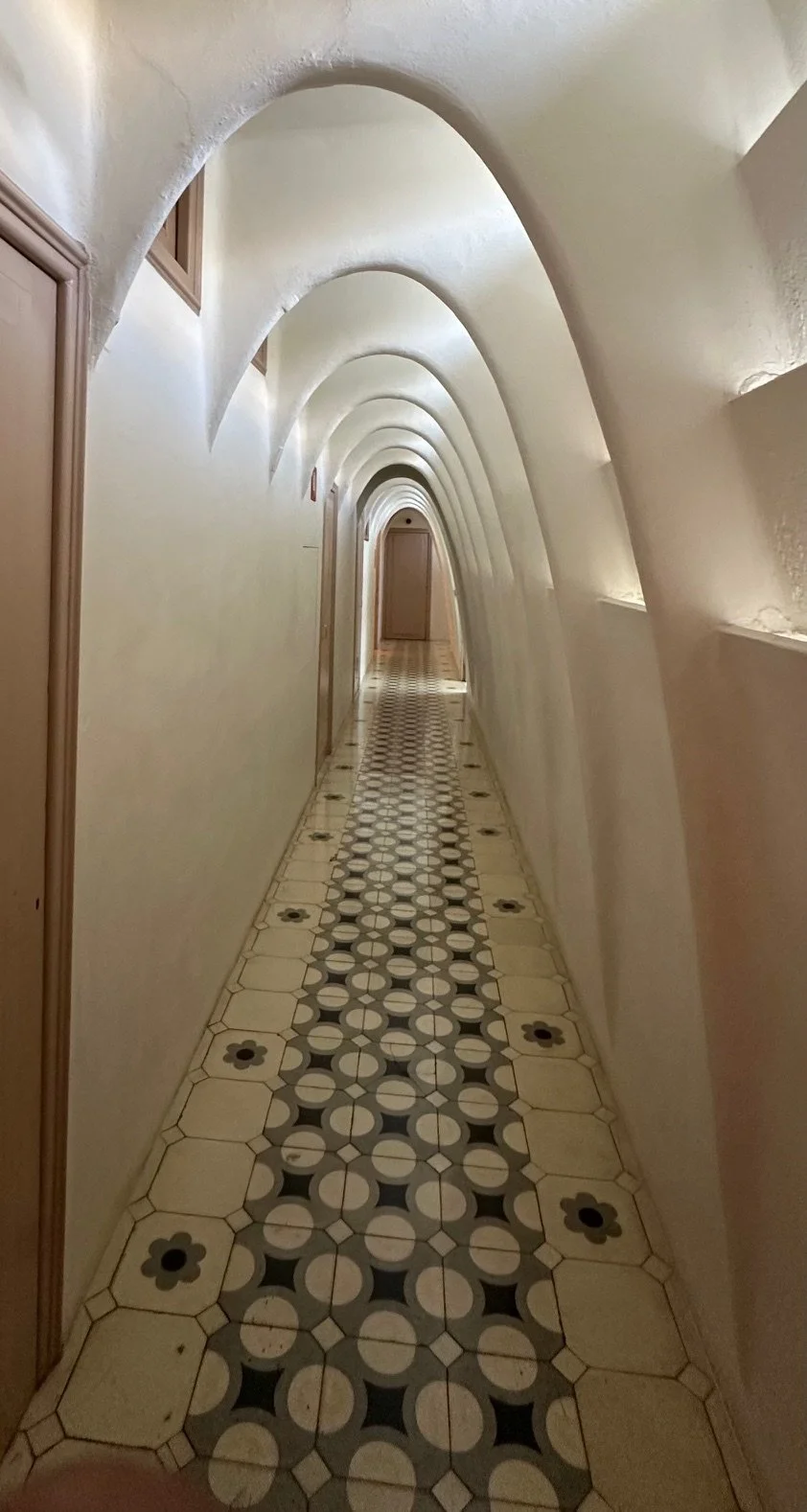 A narrow, curved hallway with a patterned tile floor, cream-colored walls, and multiple rounded archways.