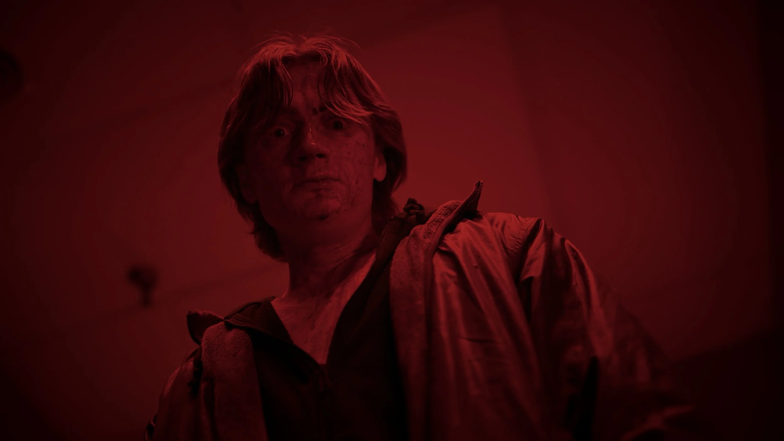 A man with disheveled hair and a distressed expression, covered in dirt or blood, in a dimly lit room with red lighting.