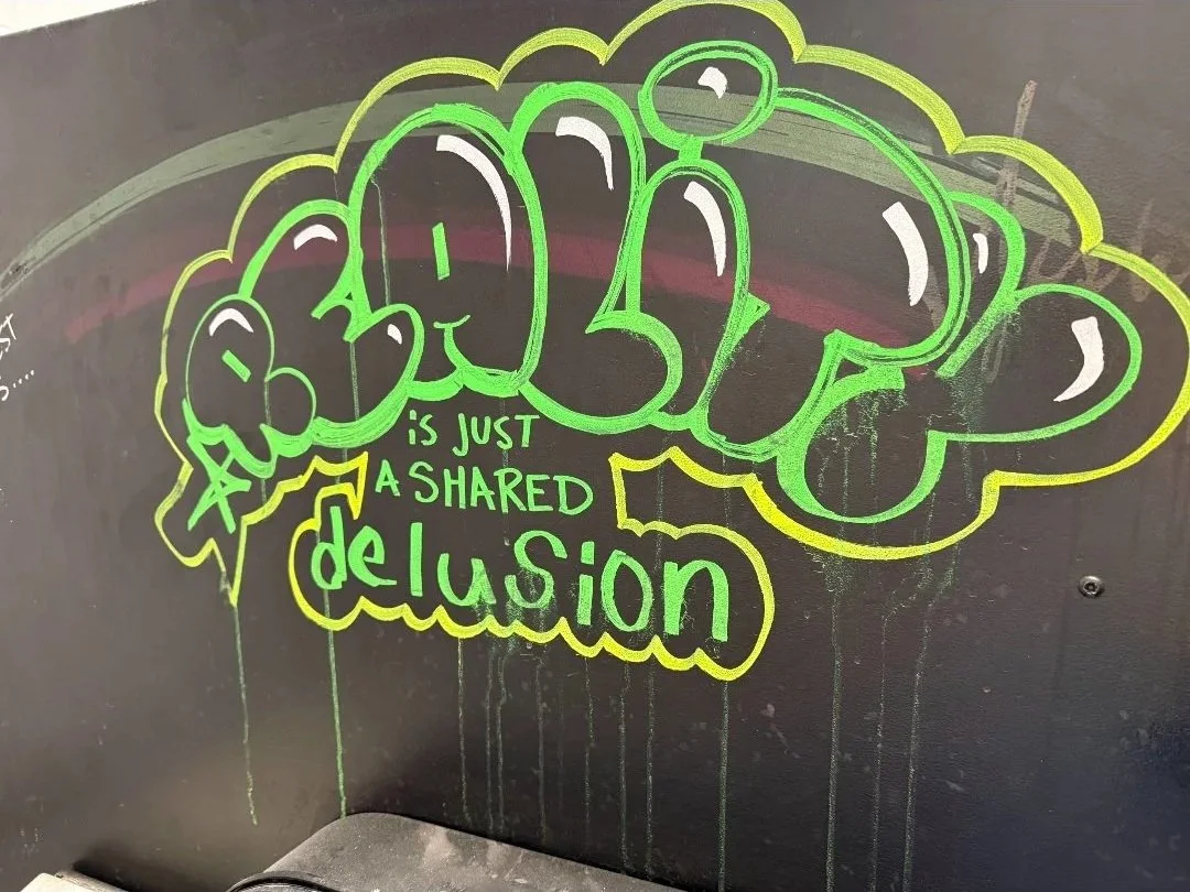 Colorful graffiti-style message on a black wall reading 'GHOUL' in large letters with the phrase 'is just a shared delusion' underneath, drawn with neon green and yellow markers.