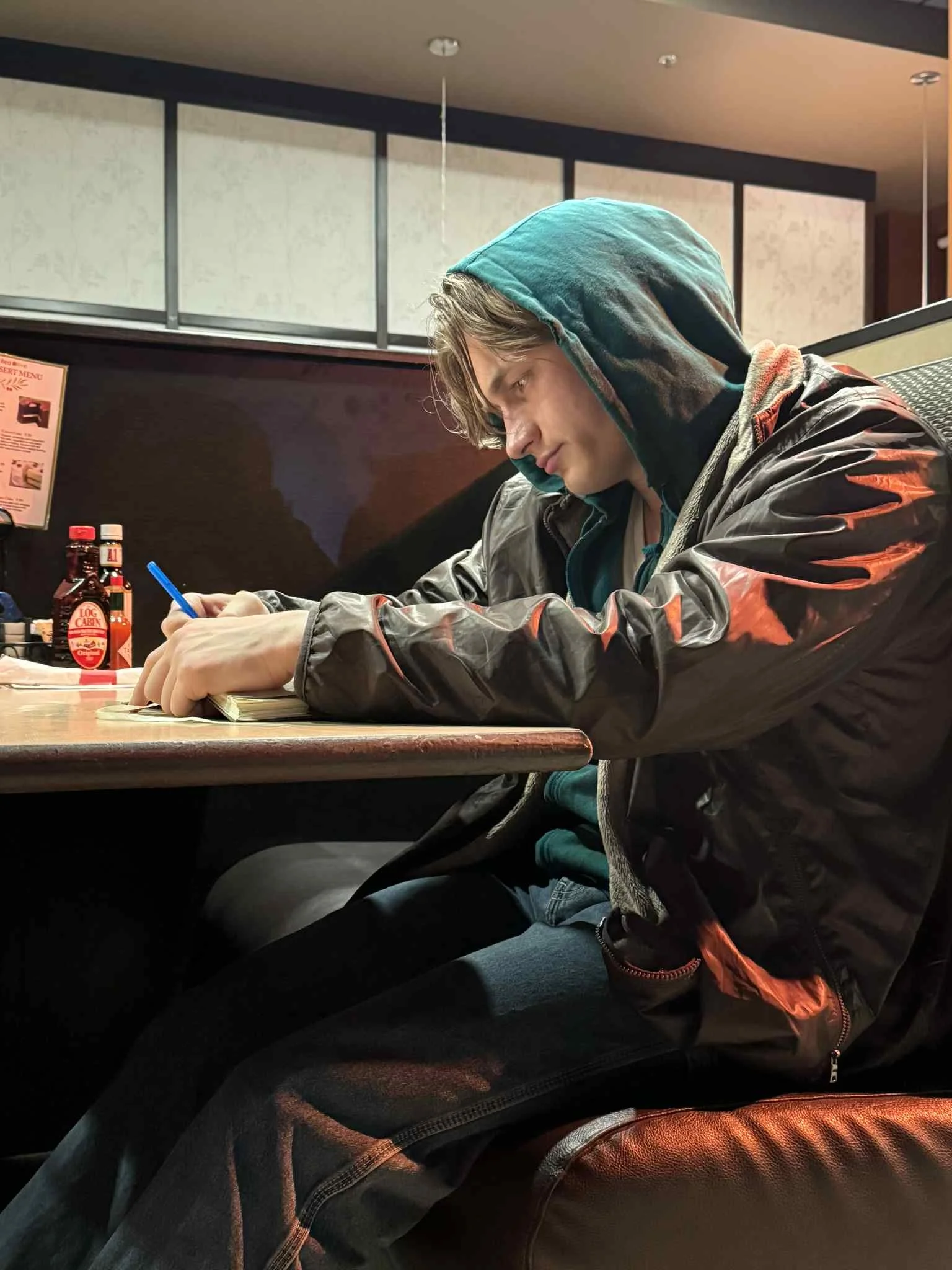 A young man in a hoodie and black jacket is sitting at a table in a restaurant, writing in a notebook with a blue pen.