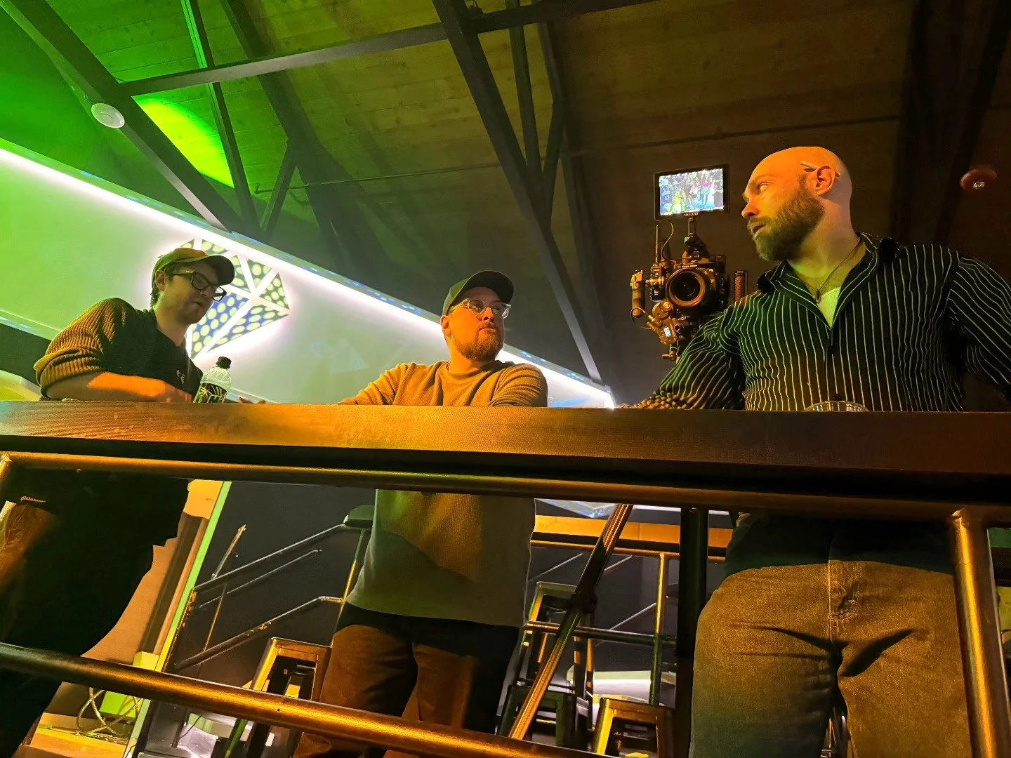 Three men on a balcony with a professional camera setup recording or streaming an event, with colorful lighting and modern decor in the background.