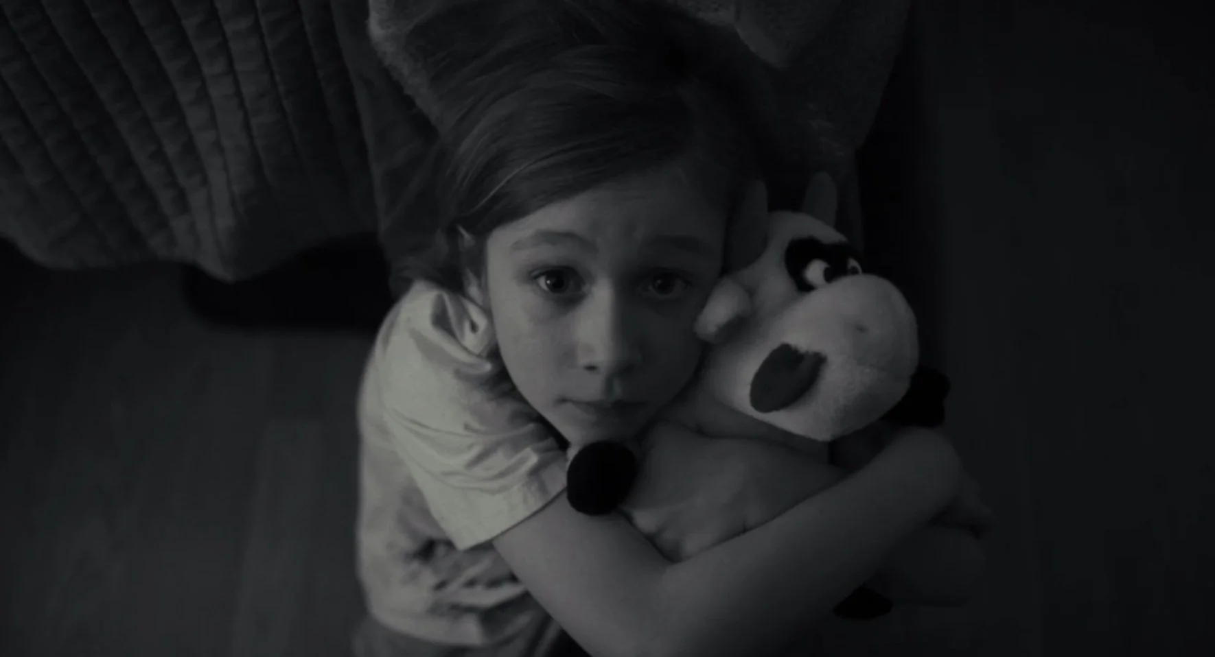 A young girl with light-colored hair hugging a stuffed Mickey Mouse toy, looking up at the camera with a somber expression in black and white.