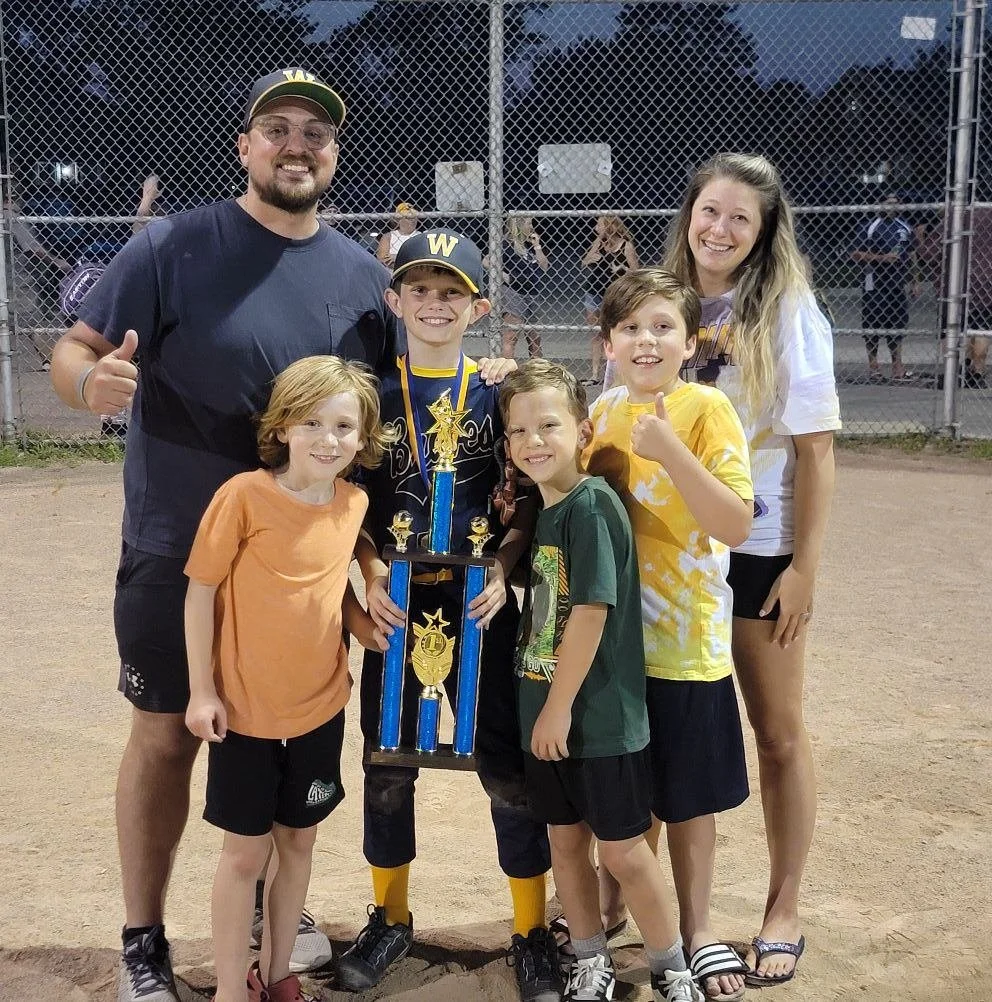 A group of six people, including four children and two adults, standing on a baseball field at night. The children are holding a large trophy, and everyone is smiling and giving a thumbs-up.