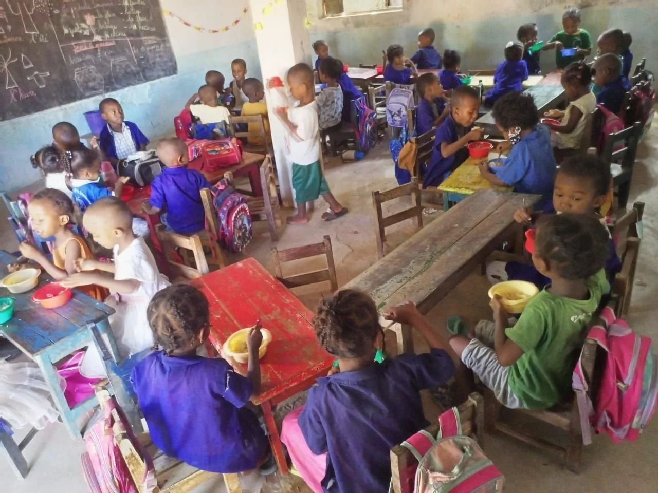 A classroom filled with young children sitting at wooden tables, eating from bowls, some with backpacks nearby, and a blackboard with diagrams on the wall.