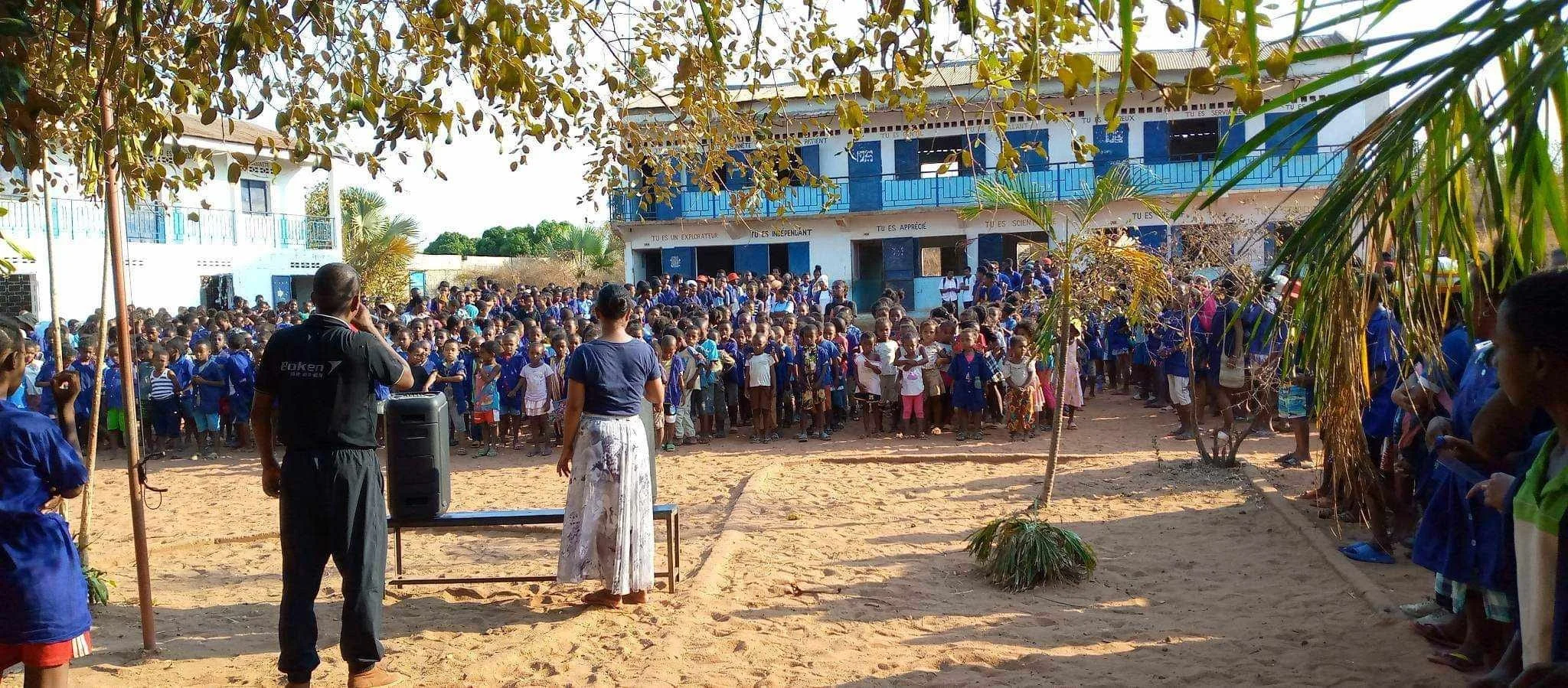 Large group of school children gathered outside school building, some in uniforms, on sandy ground, with a man and woman standing in front addressing them, and trees around.