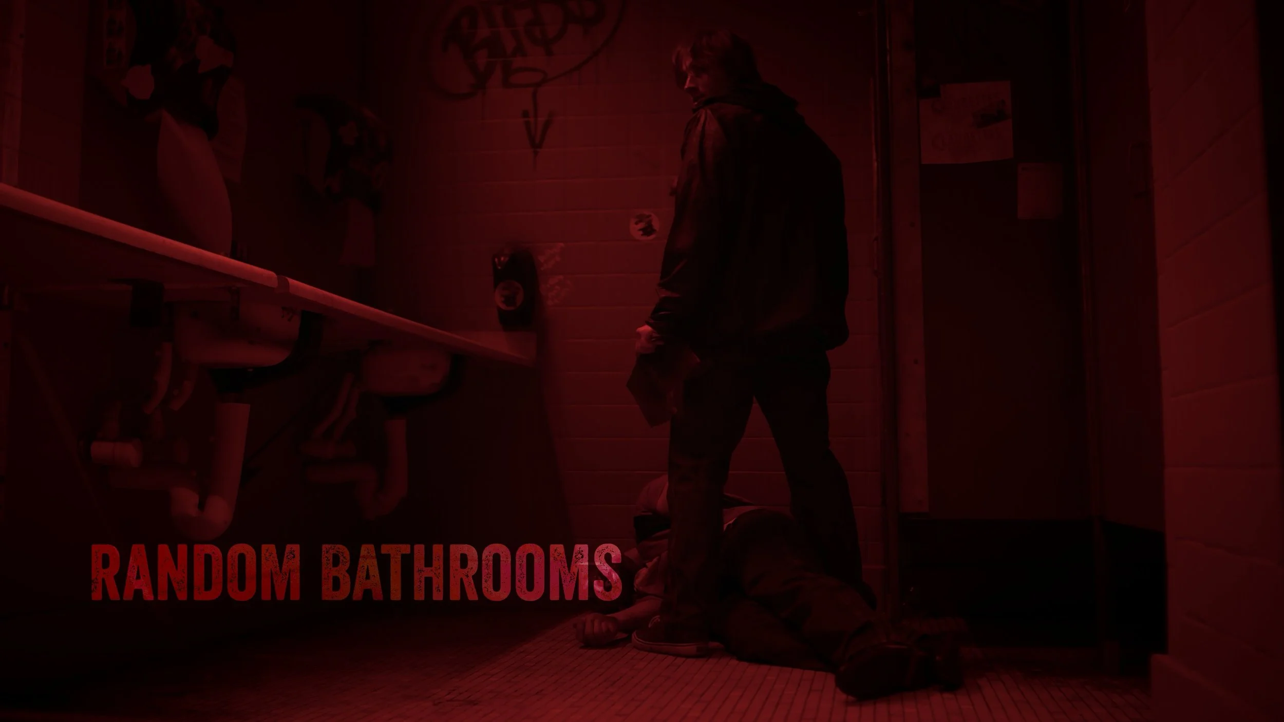 A person standing in a red-lit bathroom next to a person lying on the floor. The bathroom has sinks and graffiti on the wall with the text 'RANDOM BATHROOMS' in red at the bottom.